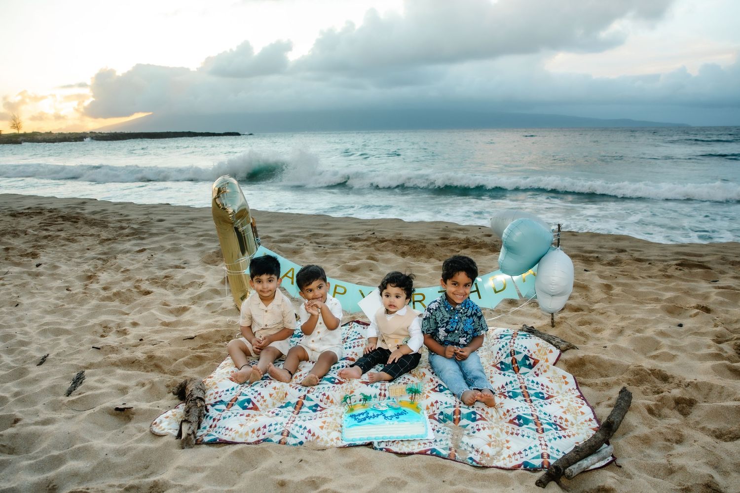 Four young children sit on a blanket on a sandy beach at sunset with balloons and a birthday cake.