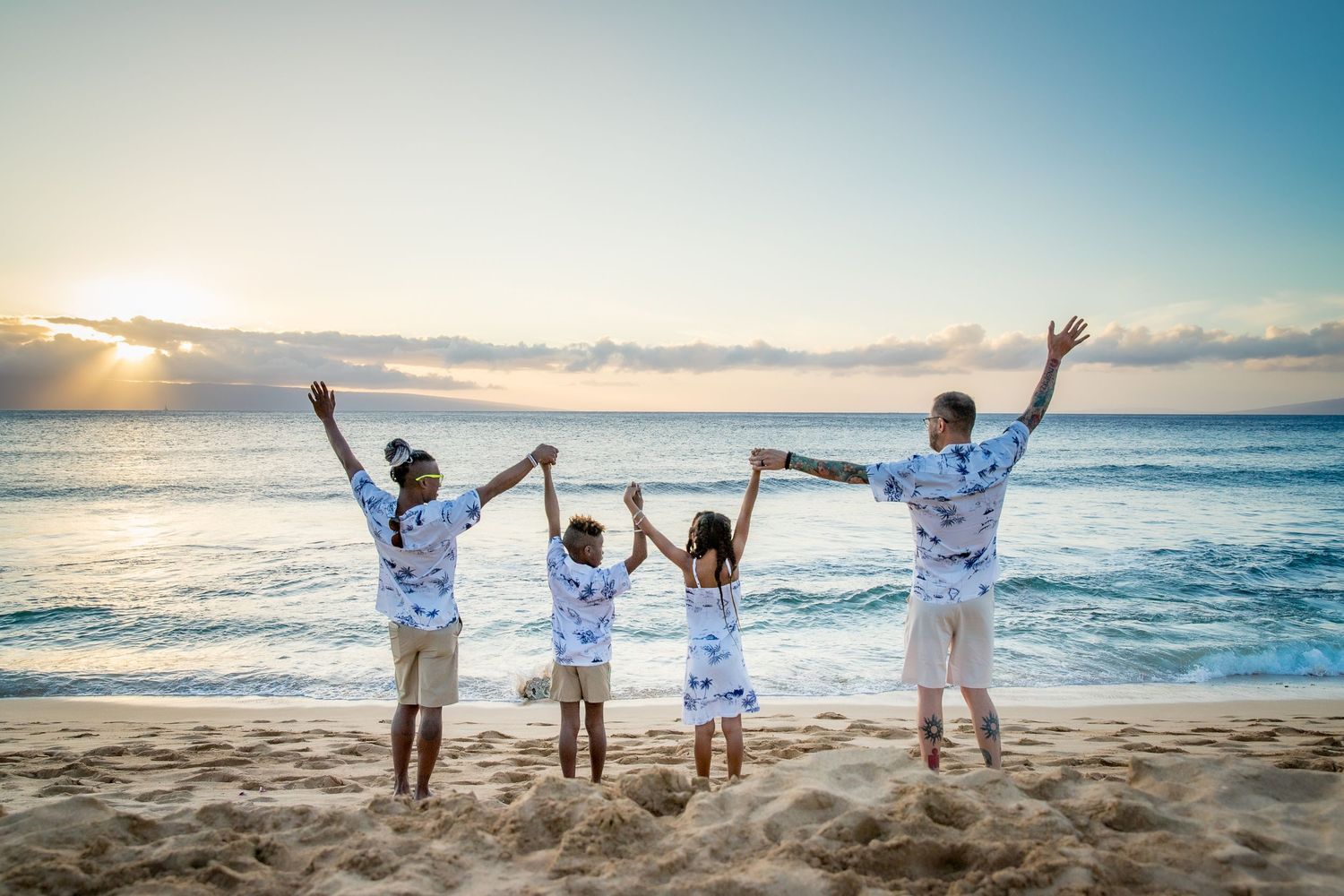 A group of people joyfully raise their arms while standing on a sandy beach at sunset.