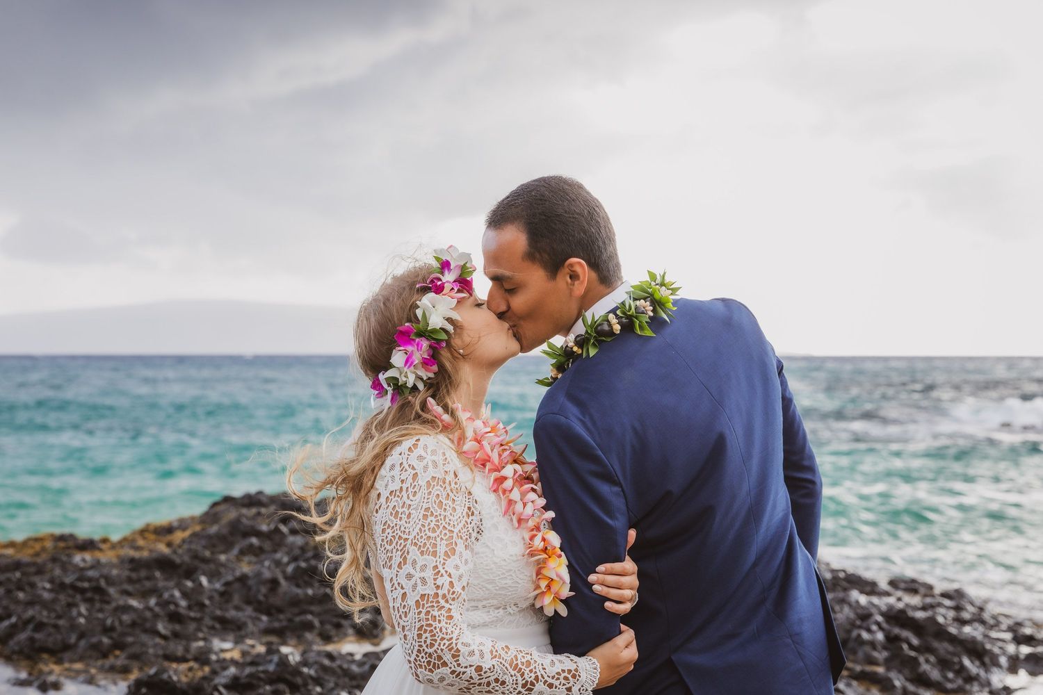 Couple in wedding attire sharing intimate moment by rocky ocean coastline.