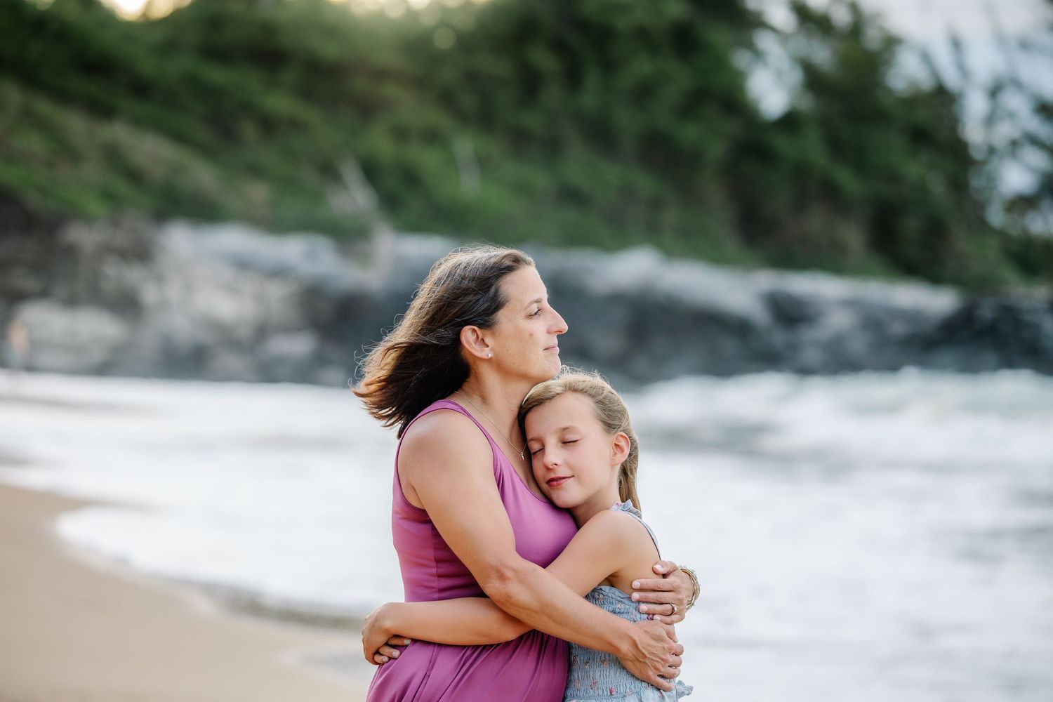 A mother and daughter share a tender hug on a sandy beach with rocky cliffs and lush greenery in the background.