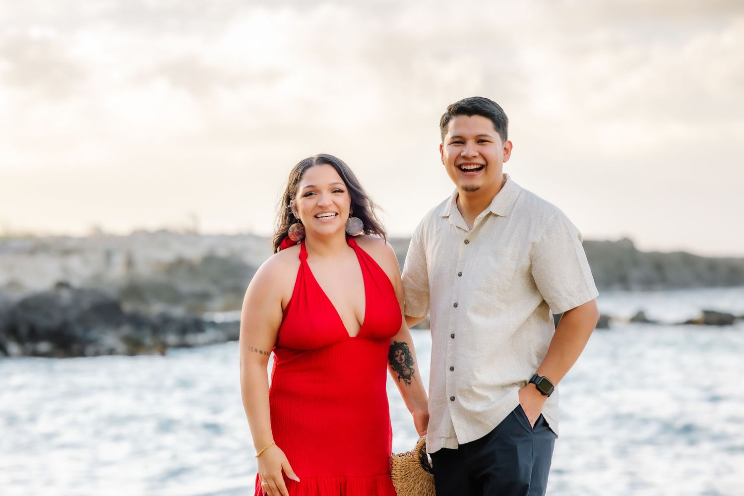 Couple smiling together on rocky Hawaiian coastline, woman in red halter dress, man in beige shirt, at golden hour.