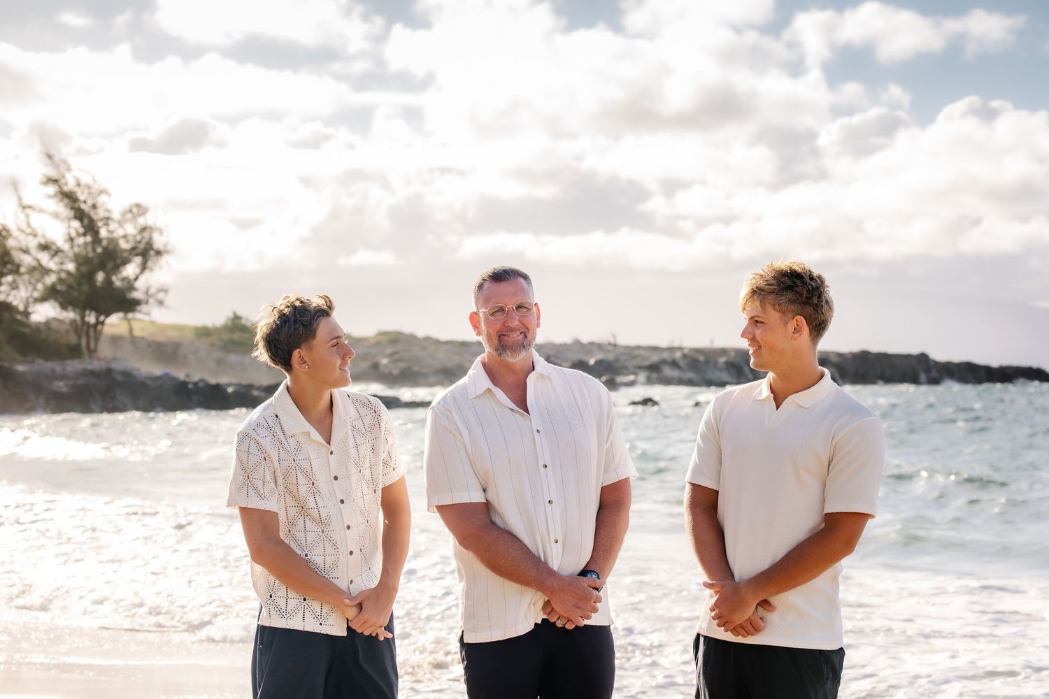 Three men smiling on a tropical beach at sunset, wearing white shirts with ocean waves in the background.