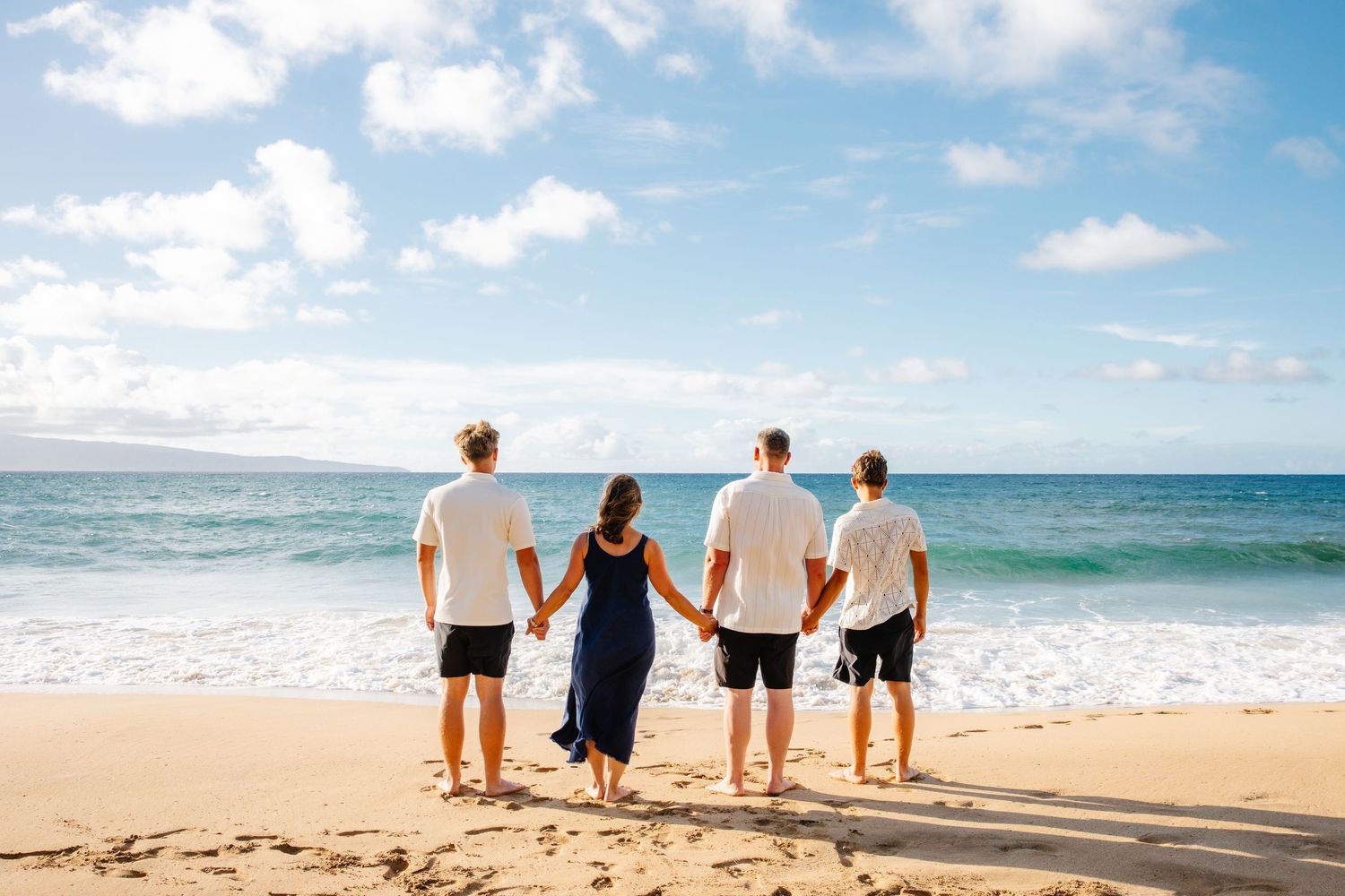 Family of four holding hands on a sandy beach, facing the ocean under a bright blue sky with white clouds.