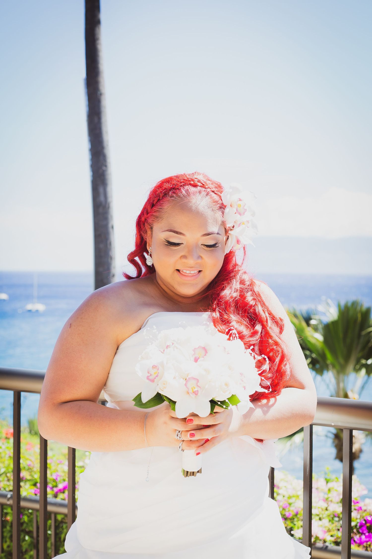 Bride with red hair holding white orchid bouquet in white strapless gown, ocean view backdrop, Maui wedding.