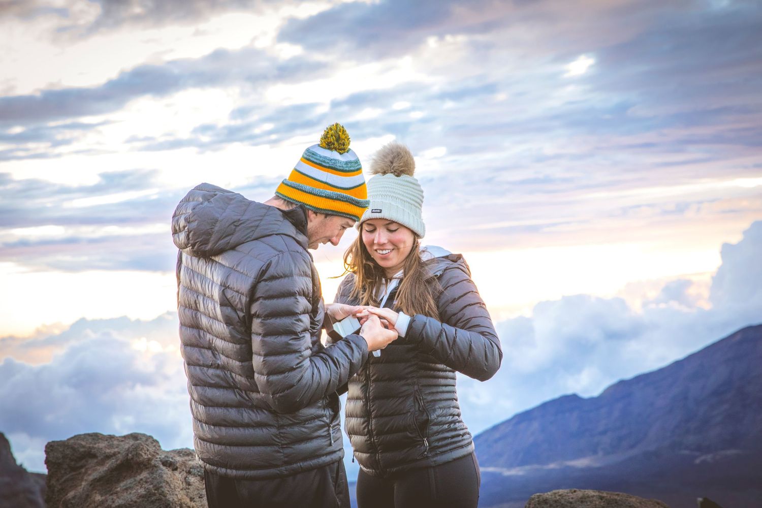 A couple in winter coats embracing against a mountain backdrop.