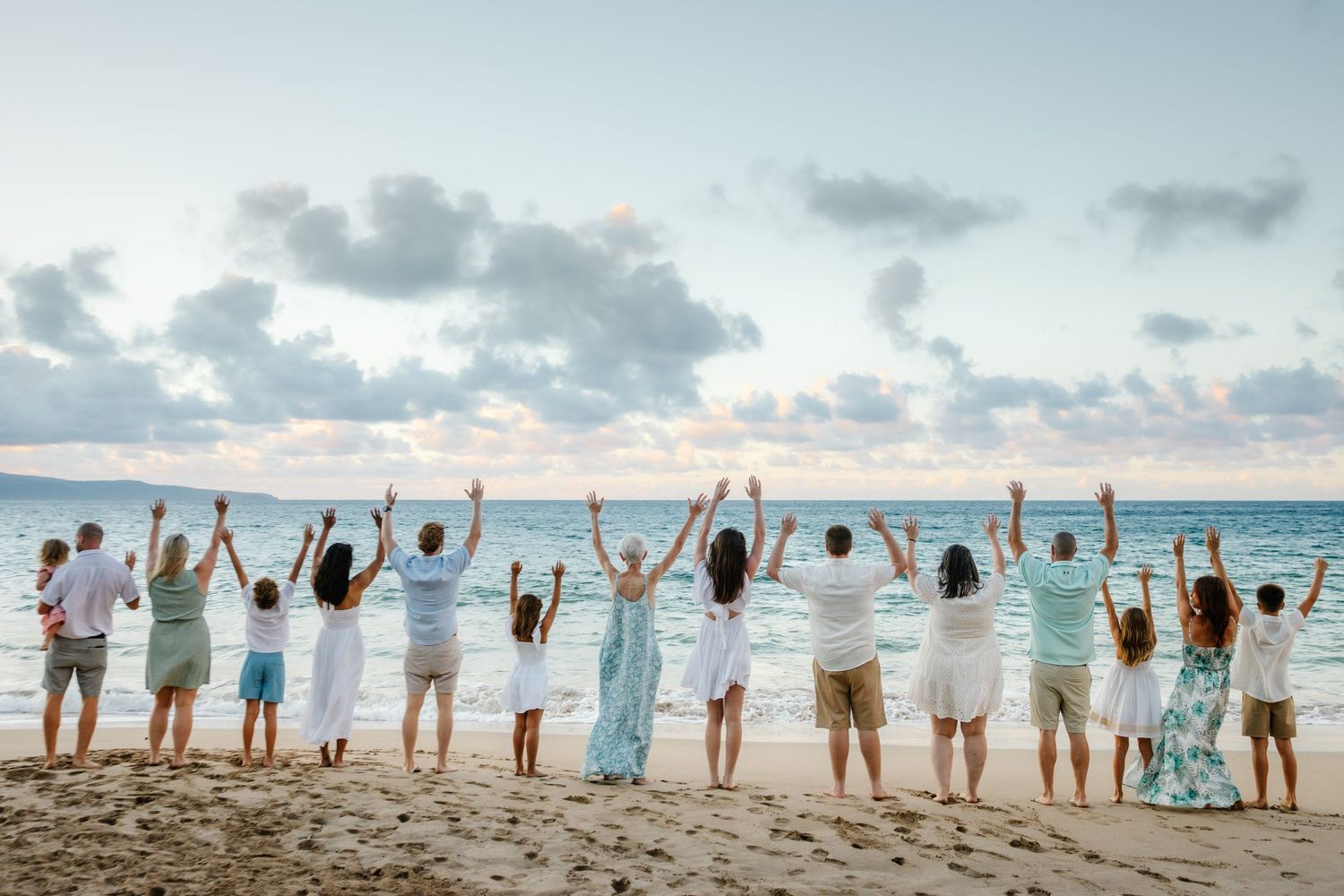 Large group of people standing on a sandy beach with arms raised, facing the ocean at sunset.