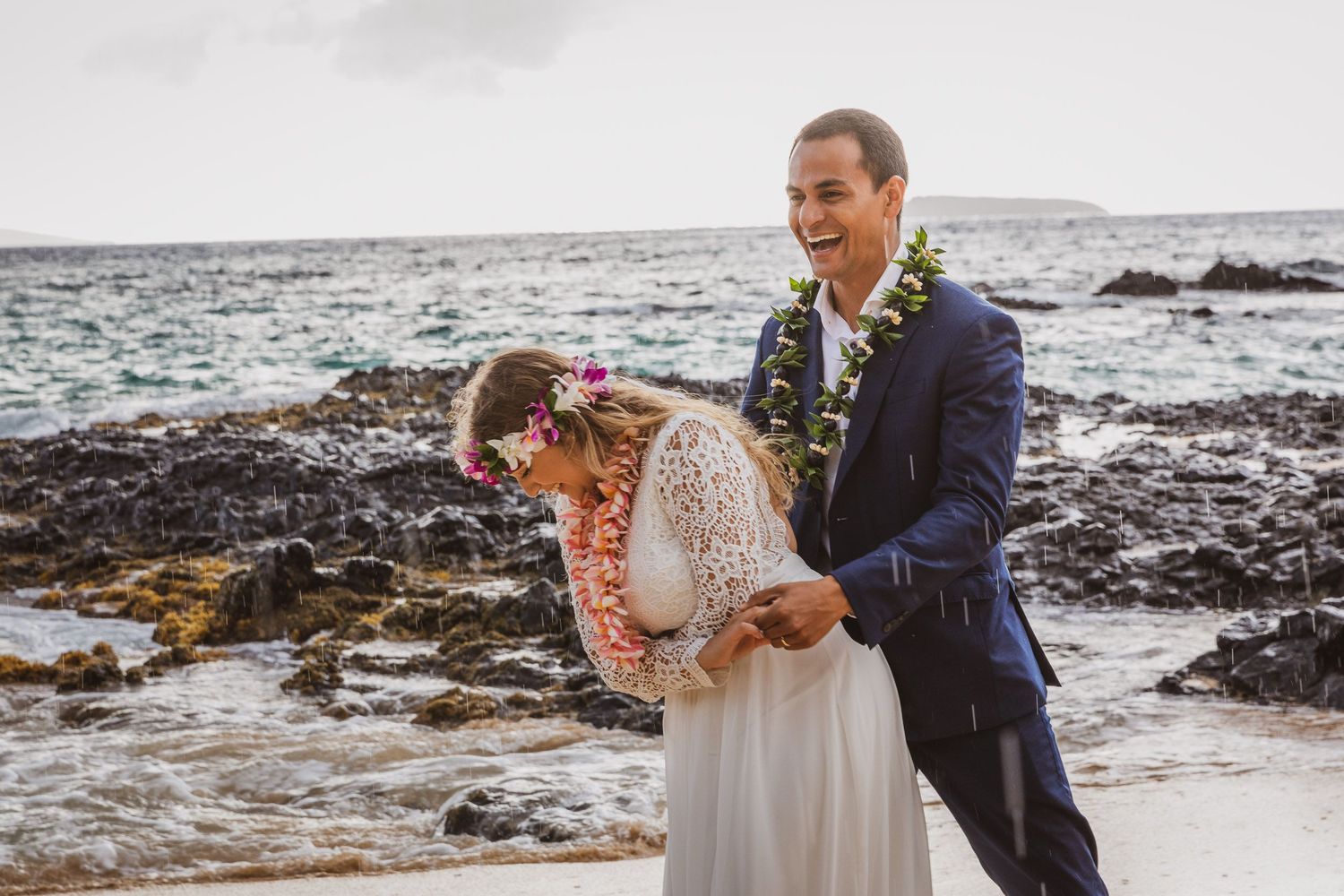Wedding couple posing on rocky beach coastline with ocean waves in background.