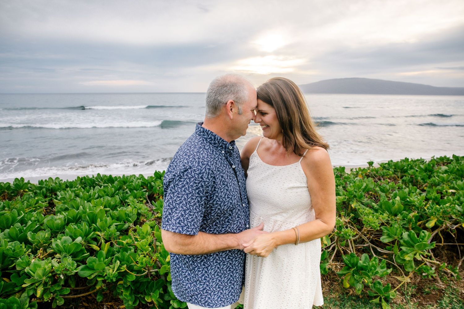 A romantic couple embraces on a tropical beach at sunset with palm trees and ocean views in Hawaii.