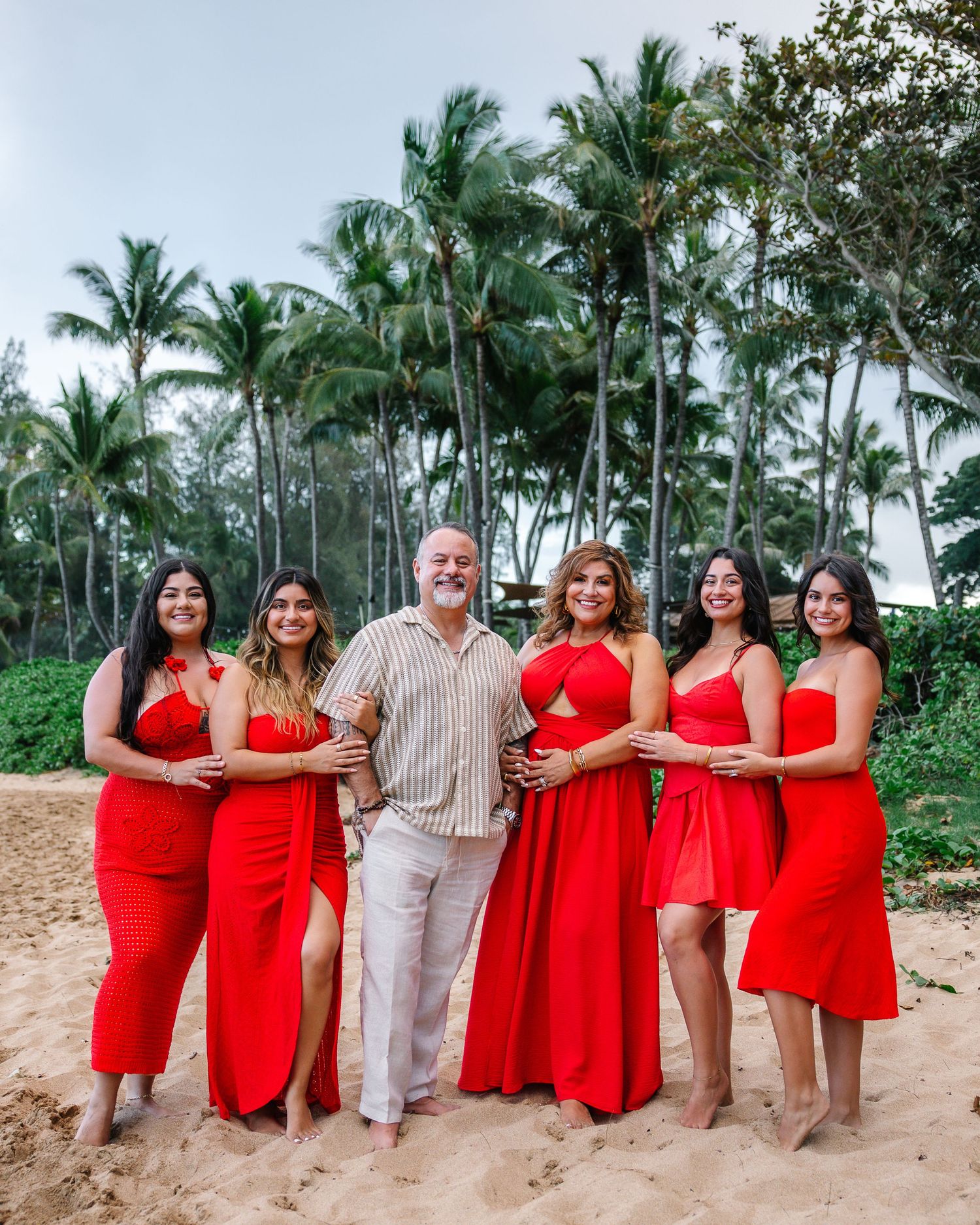 Wedding party in red dresses and beige attire poses on a tropical beach with palm trees.