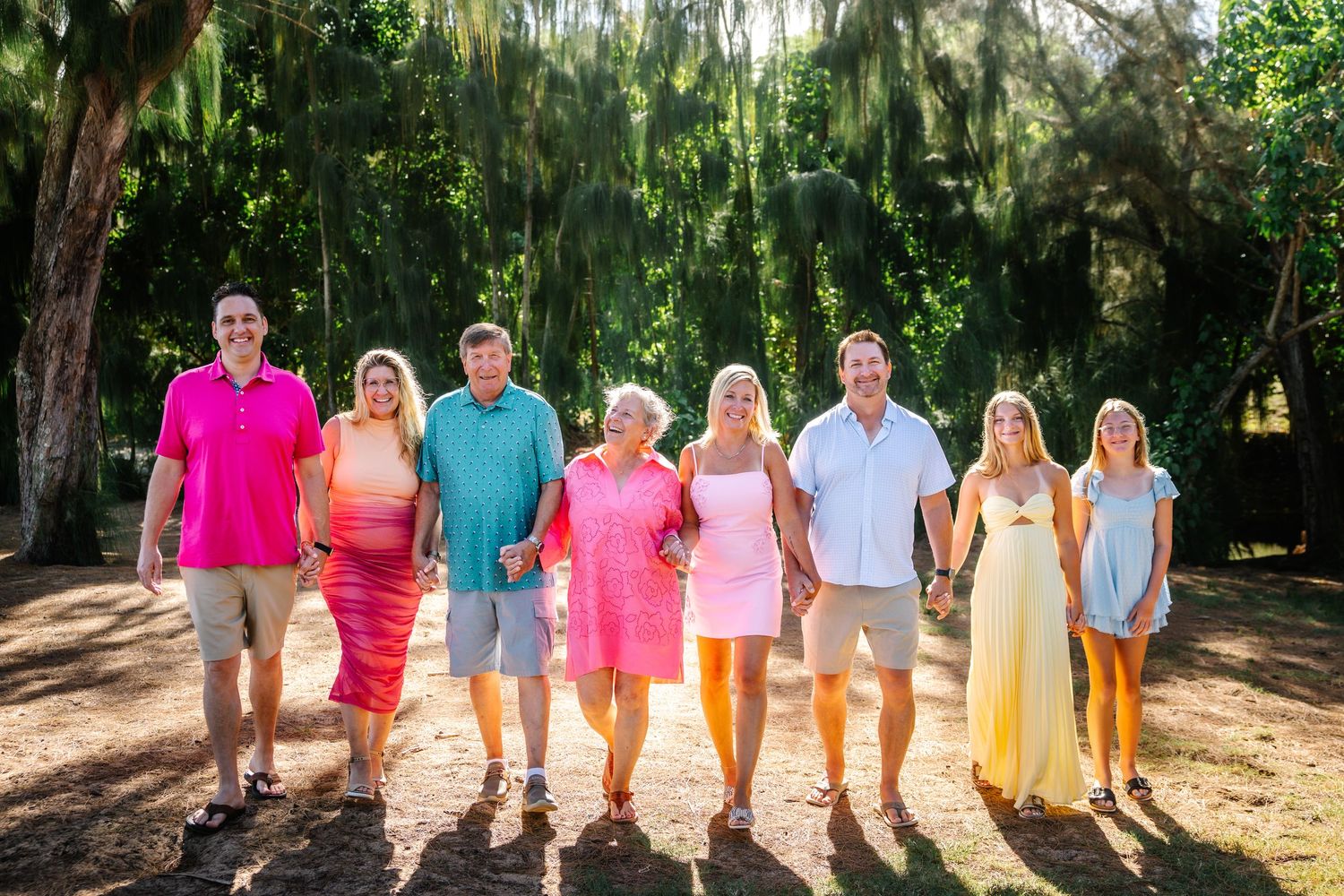 A cheerful family of seven poses outdoors in colorful outfits, surrounded by lush green trees in warm golden sunlight.
