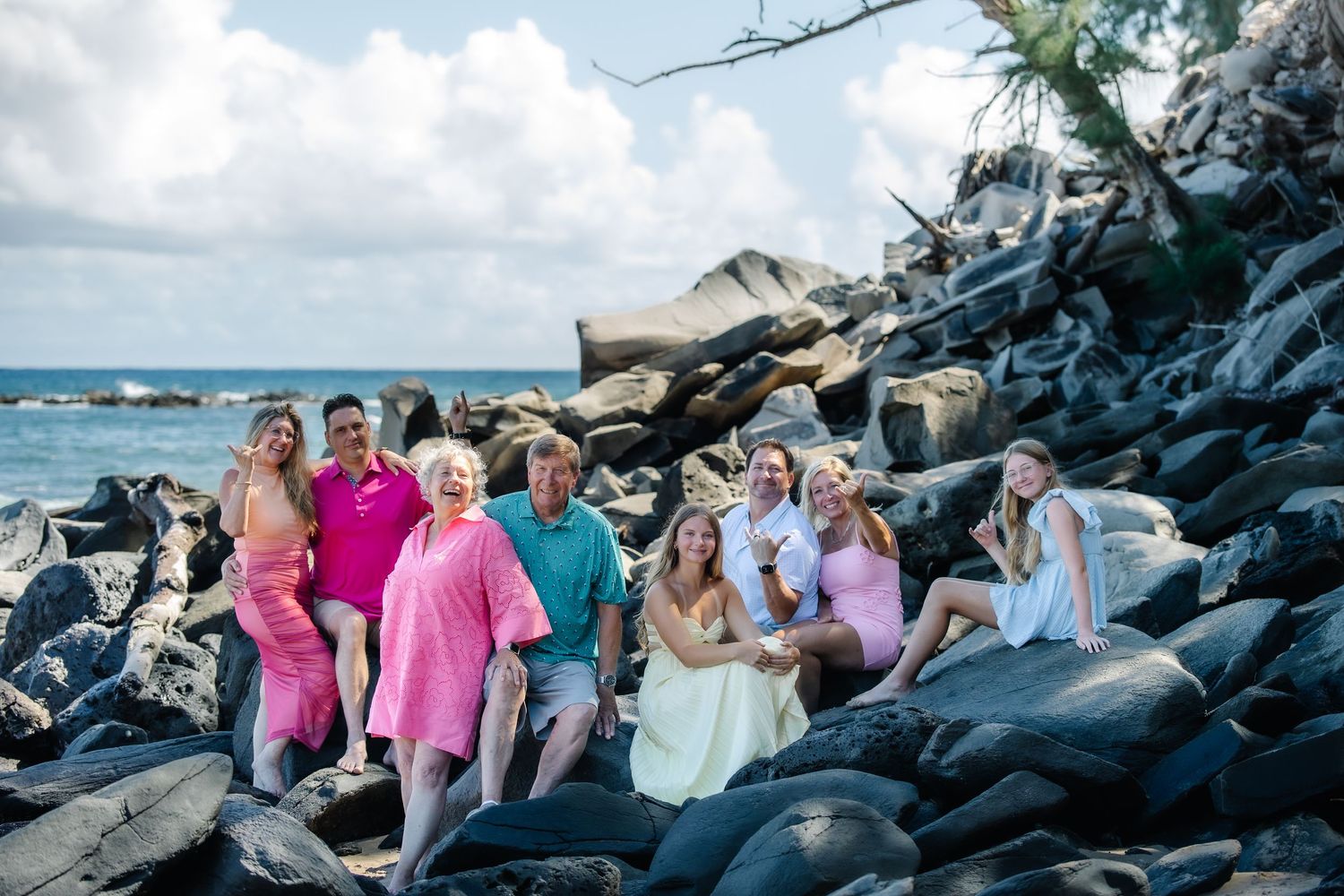 Family group photo on rocky Hawaiian beach with ocean backdrop, wearing colorful pink and teal outfits.