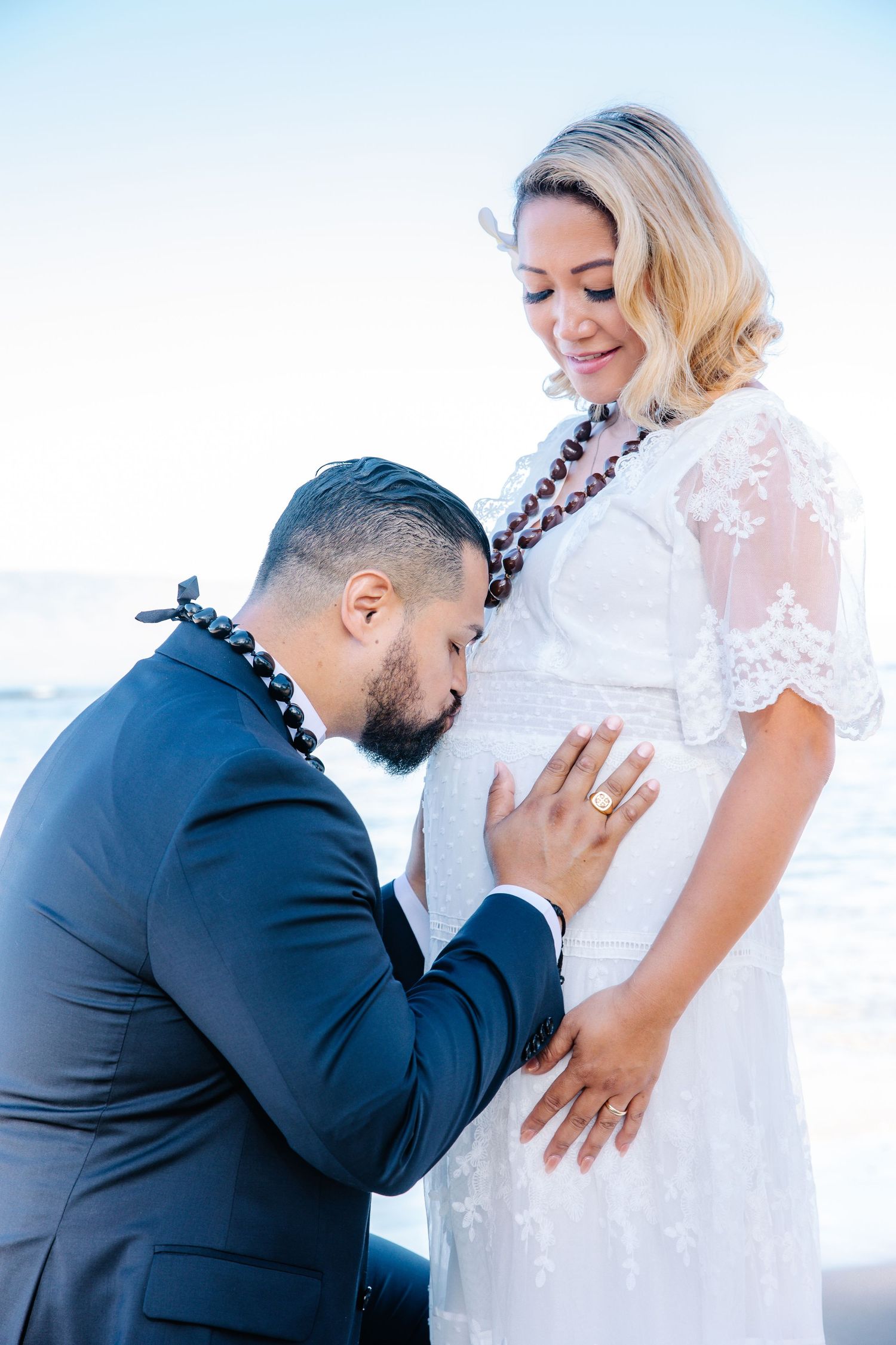 A man in a suit kisses a pregnant woman's belly during a maternity photo shoot by the ocean.