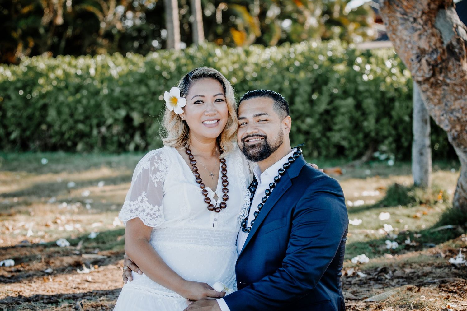 Happy couple posing outdoors, woman in white dress with flower in hair, man in navy suit, tropical garden background.