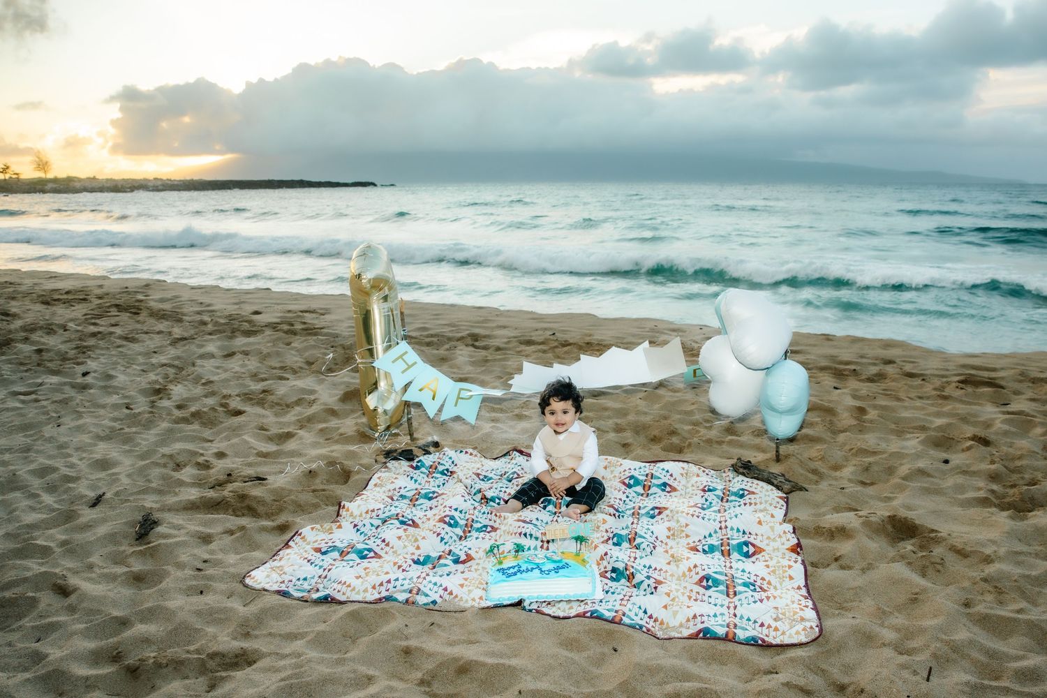 Baby sits on a quilt on a sandy beach surrounded by balloons and decorations for a first birthday celebration at sunset.