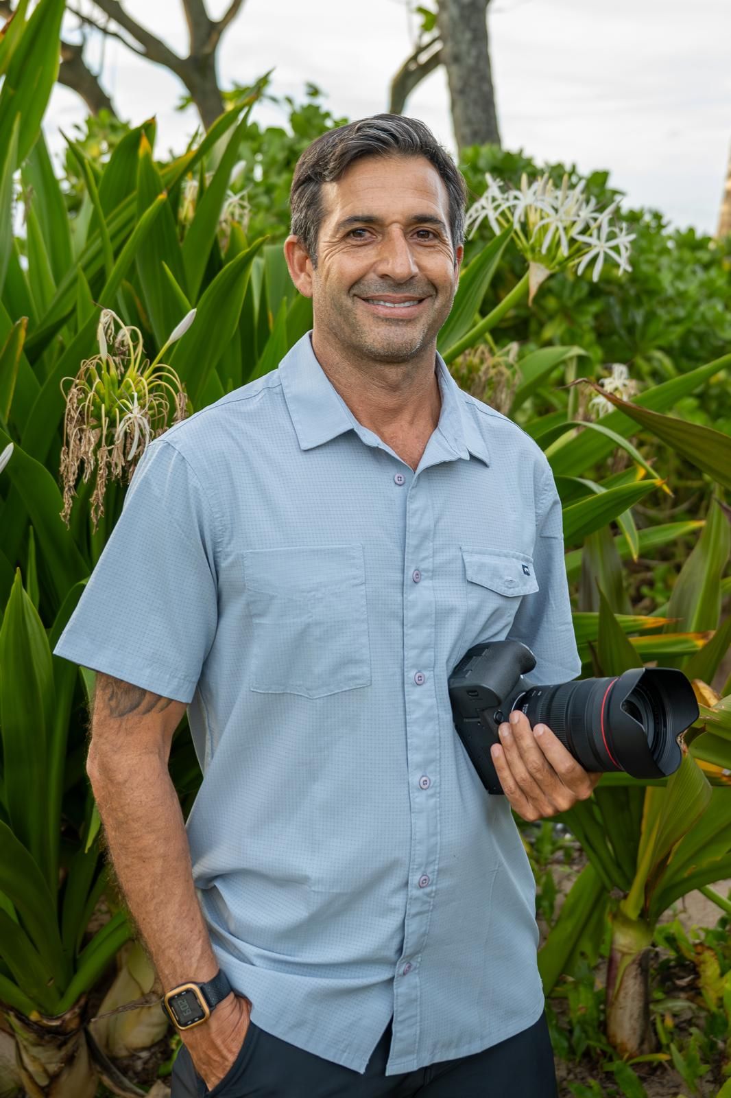 Smiling man holding a Canon camera outdoors among tropical plants, wearing a light blue shirt with a tattoo on his arm.