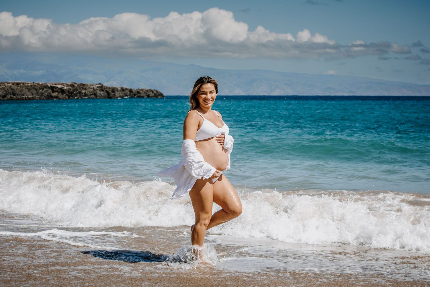 A beachside maternity photo with waves crashing and turquoise ocean waters in the background.