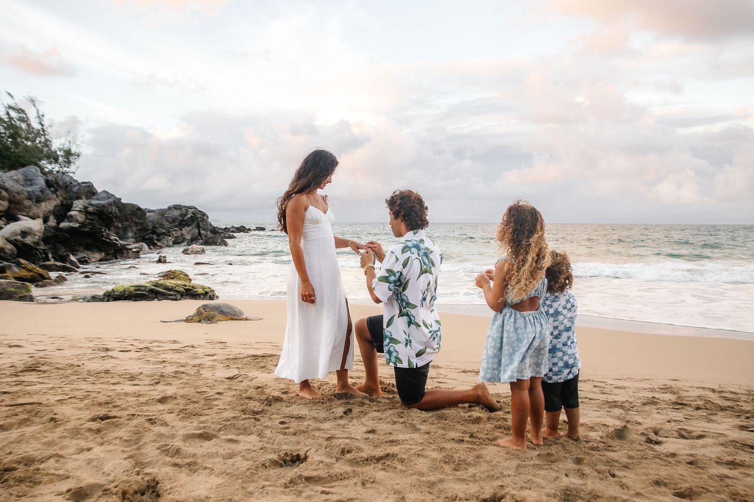 Three people in white and floral outfits share a tender moment on a sandy Hawaiian beach at sunset.