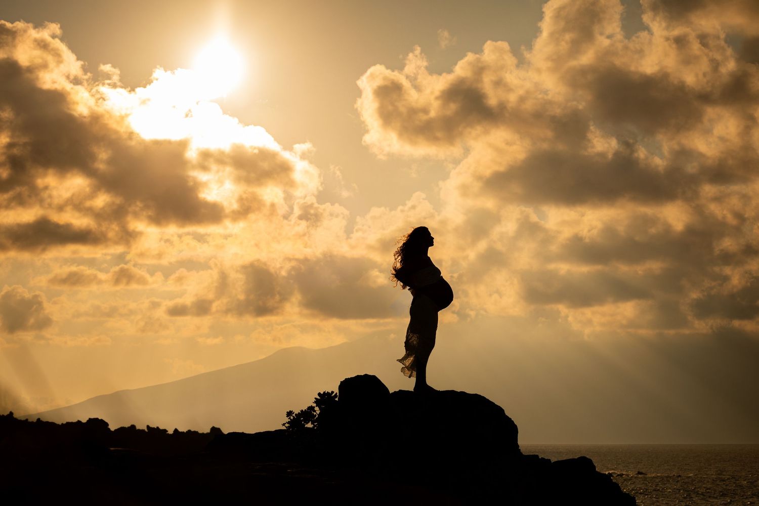 Silhouette of a woman standing on rocks at sunset with dramatic golden clouds over the ocean.