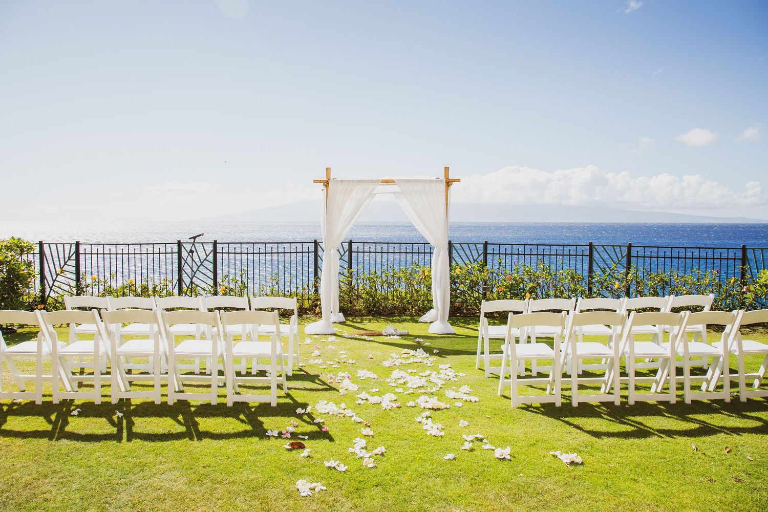 Outdoor wedding ceremony setup with white chairs, floral arch, and ocean view on a sunny day.