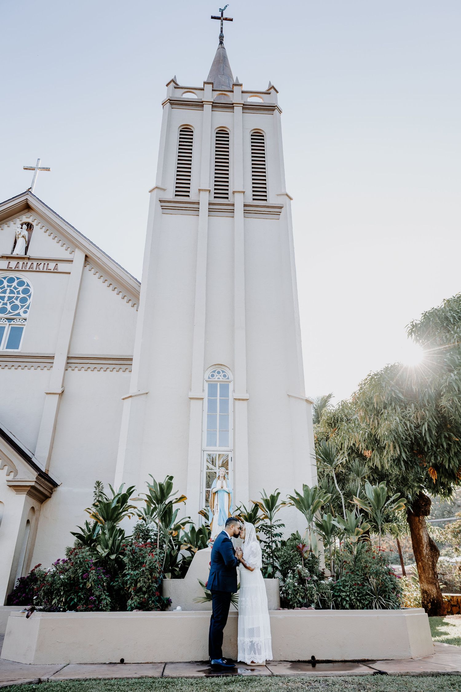 Couple sharing a romantic moment in front of a white church tower with lush tropical greenery on their wedding day.