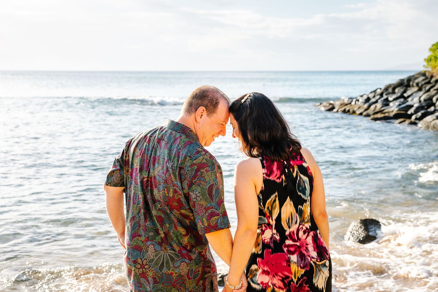 A couple in colorful attire walks along a rocky Hawaiian coastline.