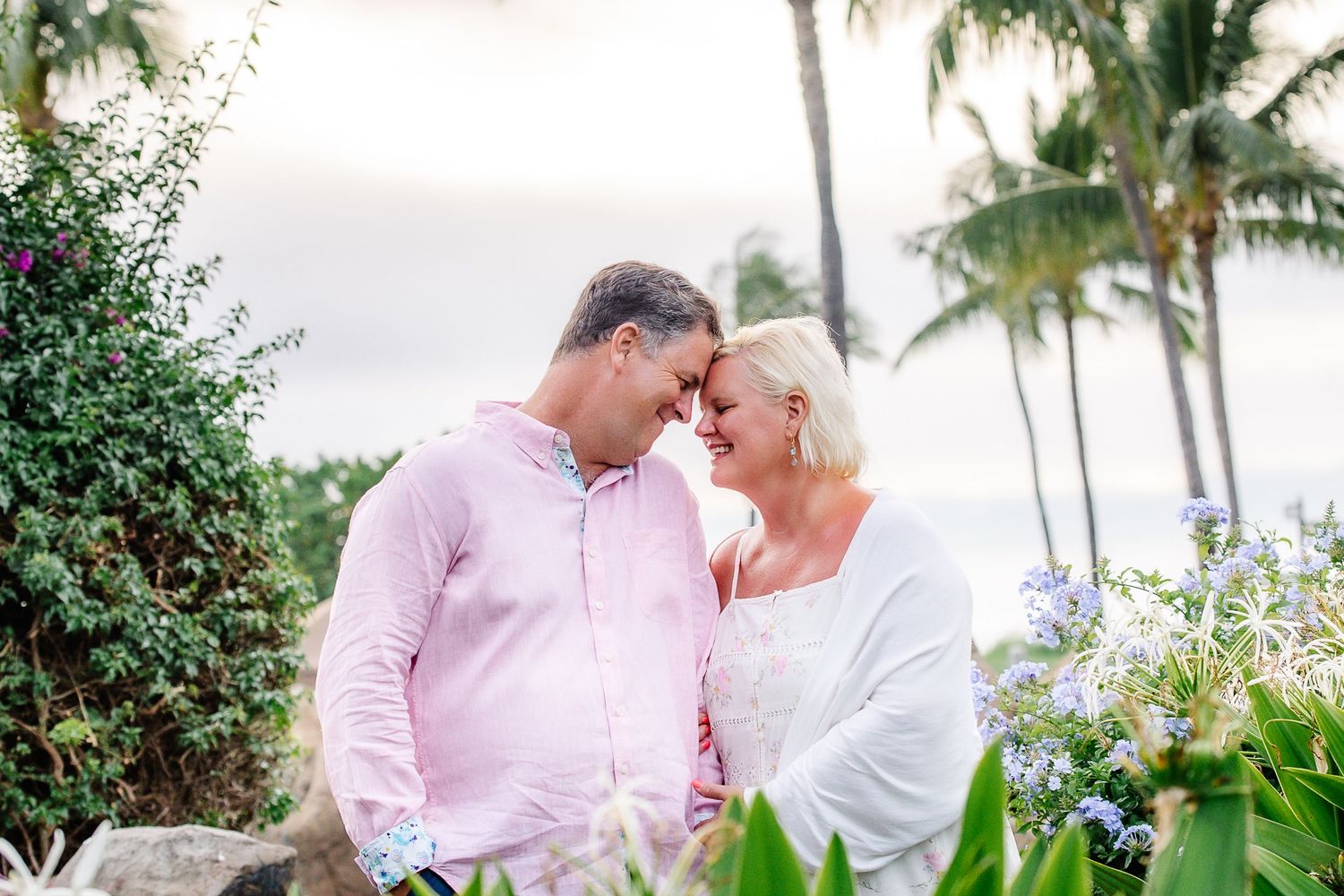 Romantic couple touching foreheads and smiling outdoors among tropical palms and colorful flowers.