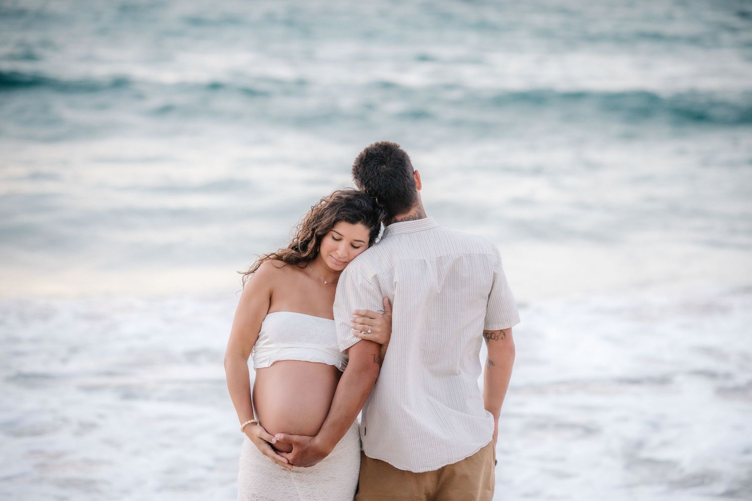 Pregnant couple embracing on a beach, woman in white dress cradling baby bump with ocean waves behind them.