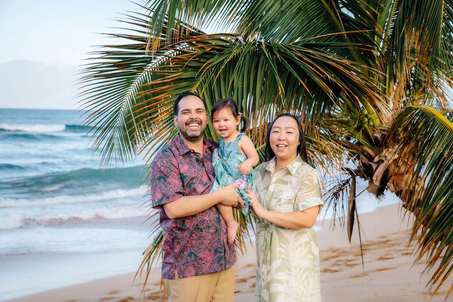 A couple poses together on a tropical beach with palm trees and ocean waves in the background.