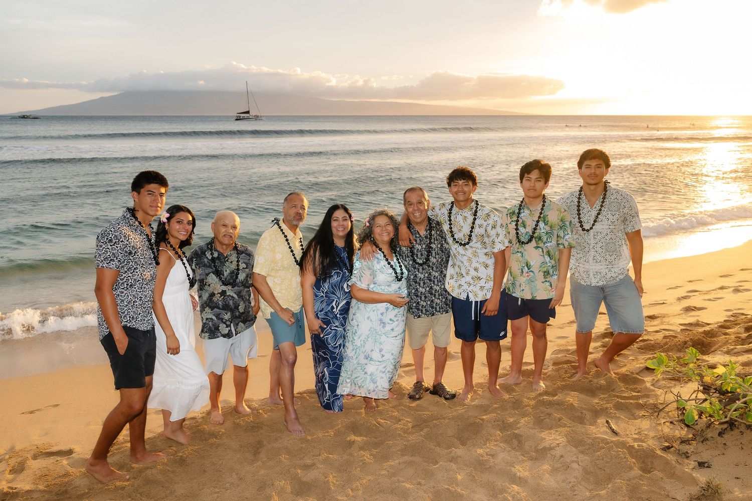 A group of people wearing Hawaiian leis stand together on a beach at sunset with ocean and mountains in the background.