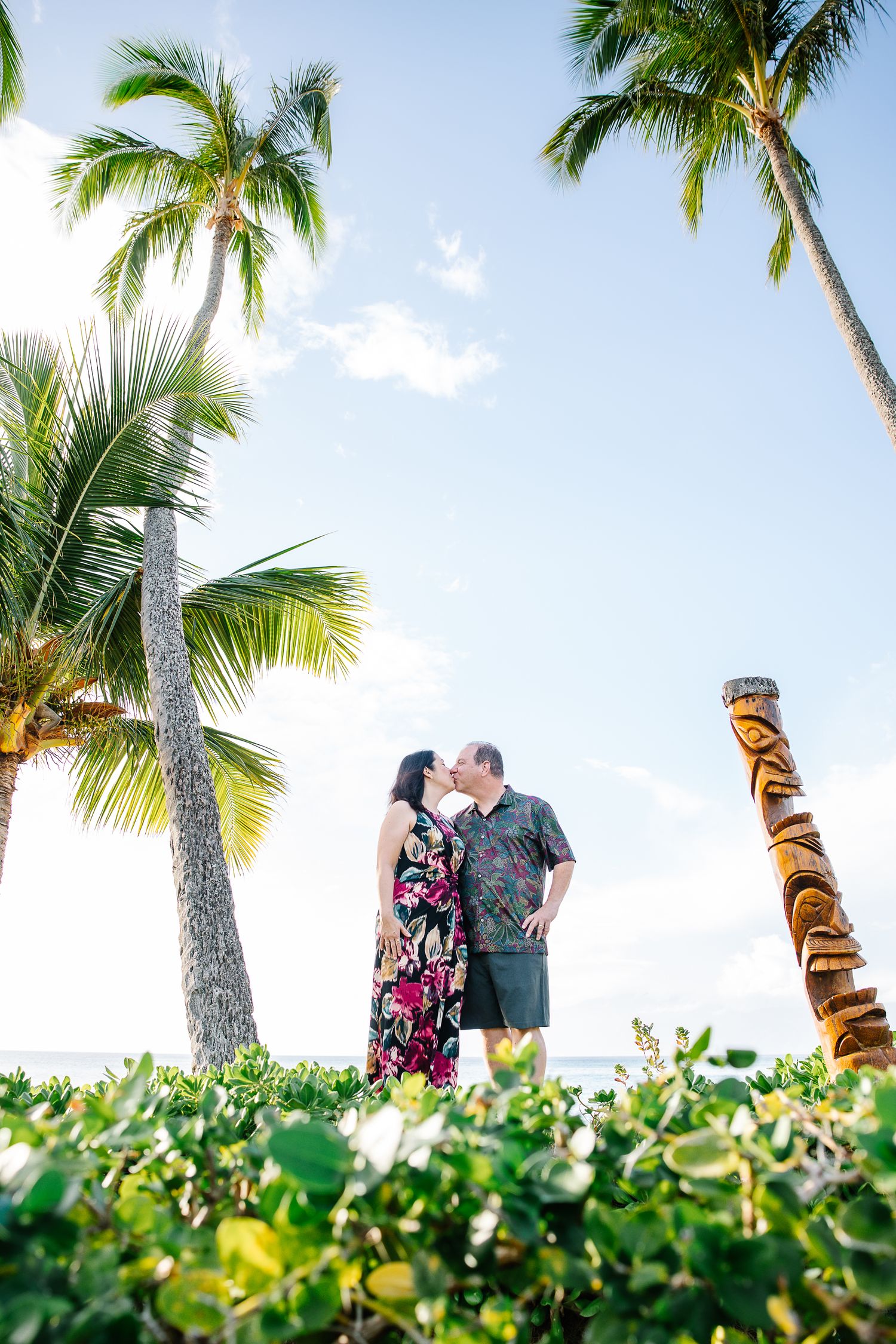 A couple walks along a tropical path lined with palm trees and tiki statues.