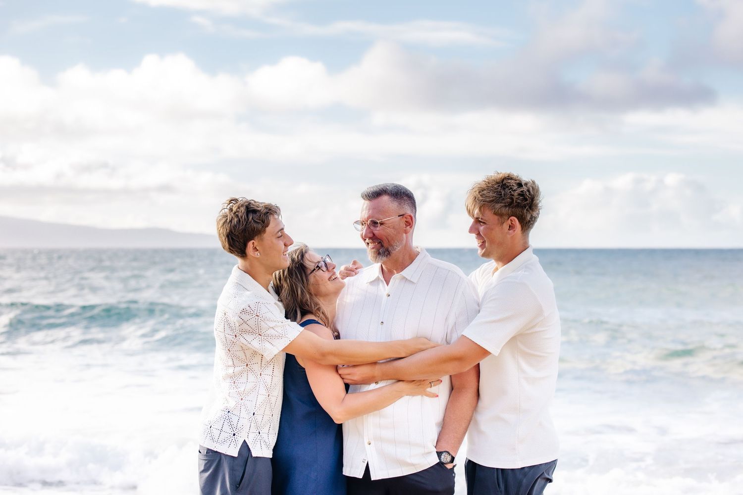 Family of four laughing and embracing on a beautiful Hawaiian beach with ocean waves and cloudy sky behind them.
