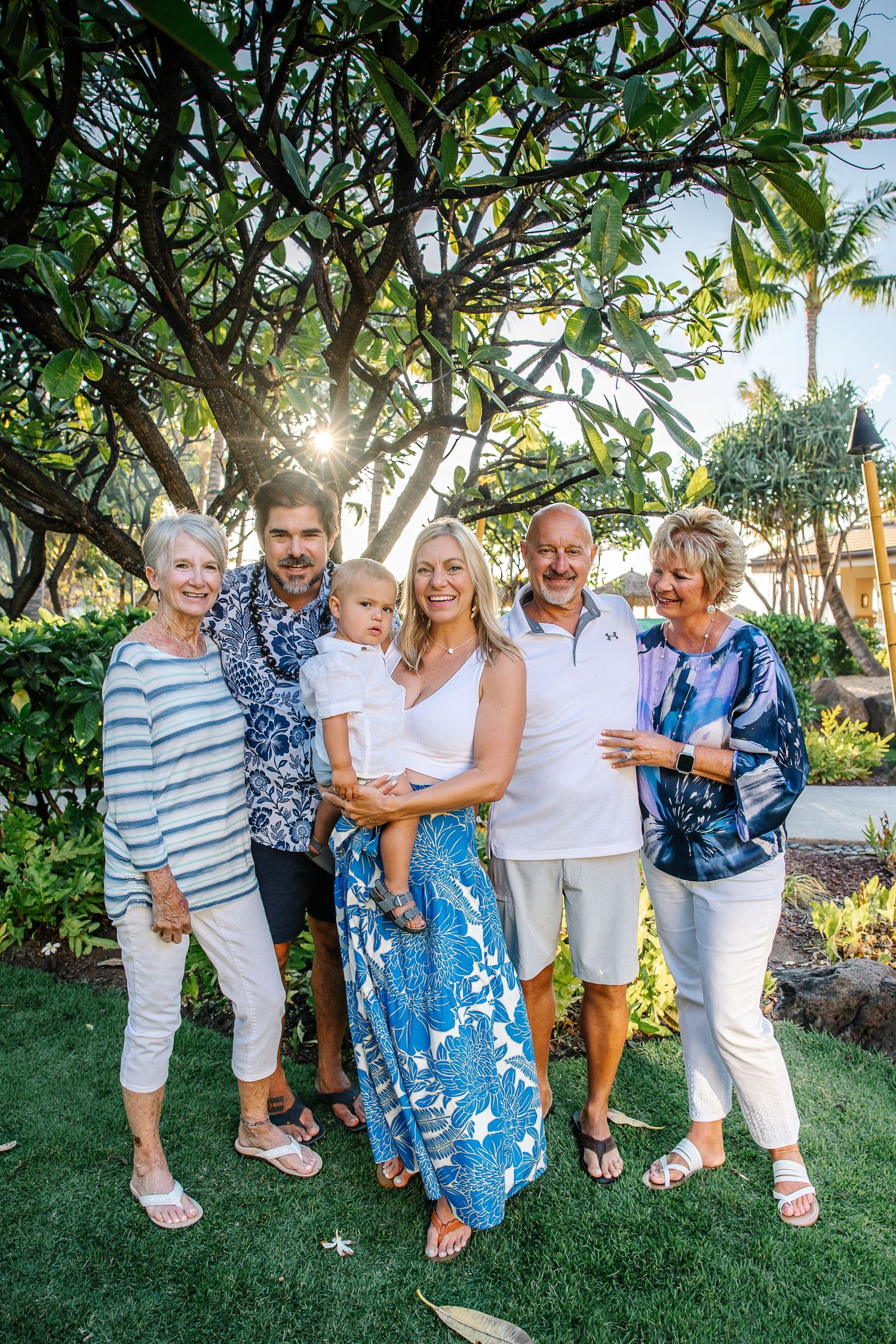Extended family gathers for a group photo on a lush Hawaiian lawn with palm trees and ocean views.