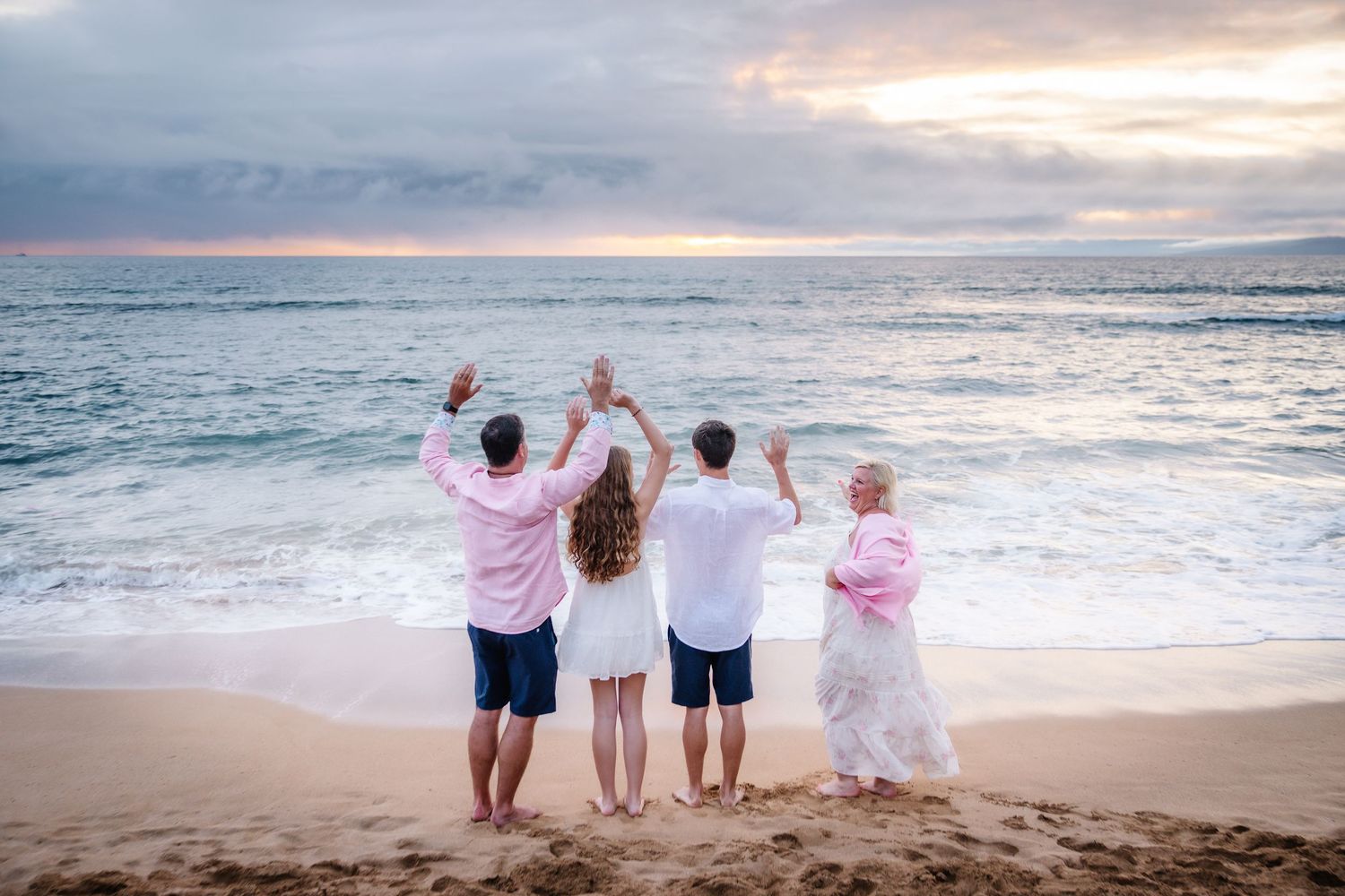 Family of four celebrating on a sandy beach at sunset, raising hands together facing the ocean waves.