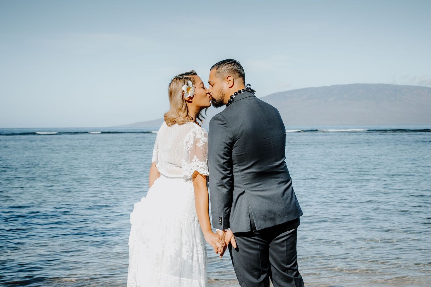 Newlywed couple kissing on a tropical beach in Maui, bride in white lace dress, groom in dark suit.