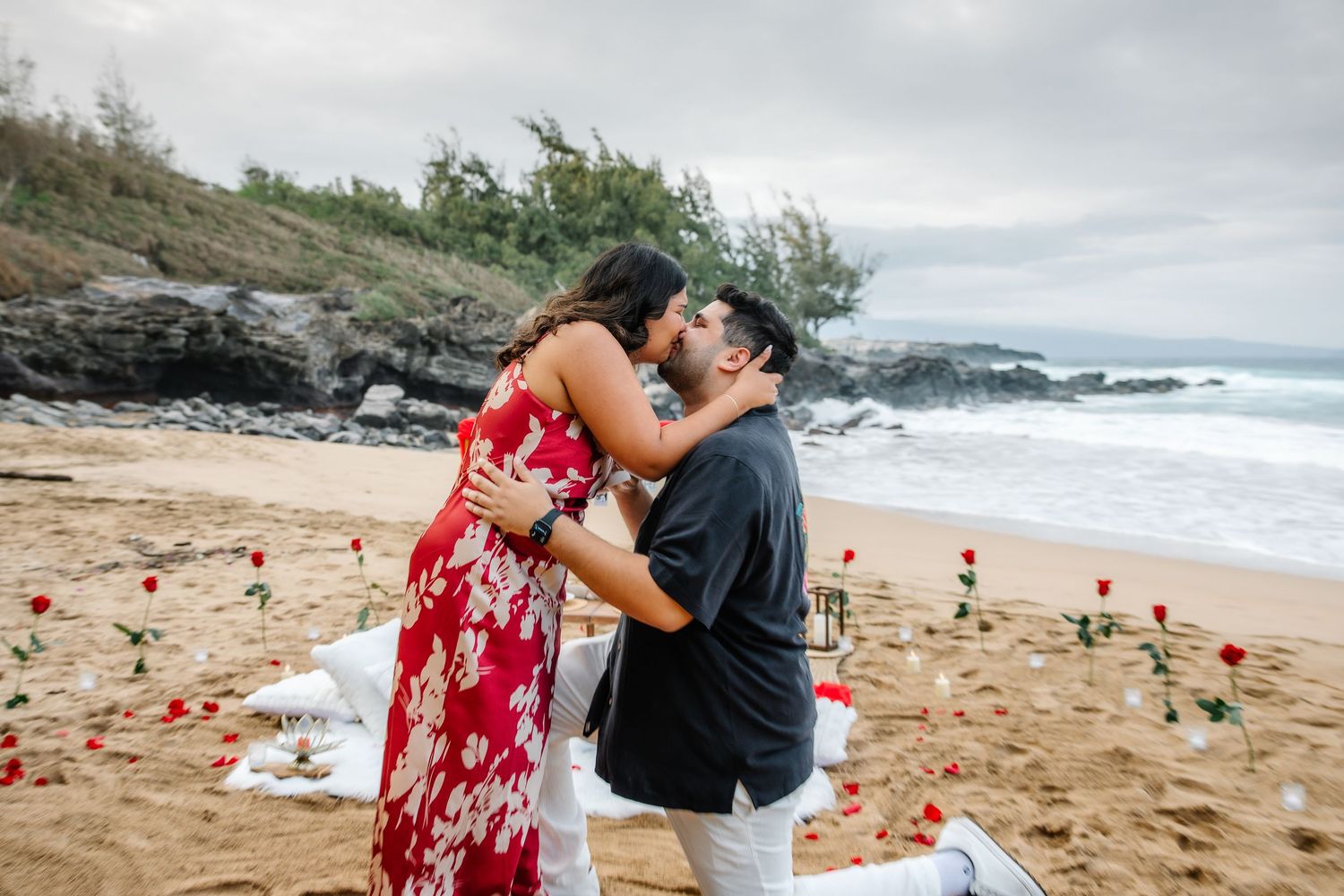 A romantic beach proposal setup with red rose petals scattered on the sand near ocean cliffs.