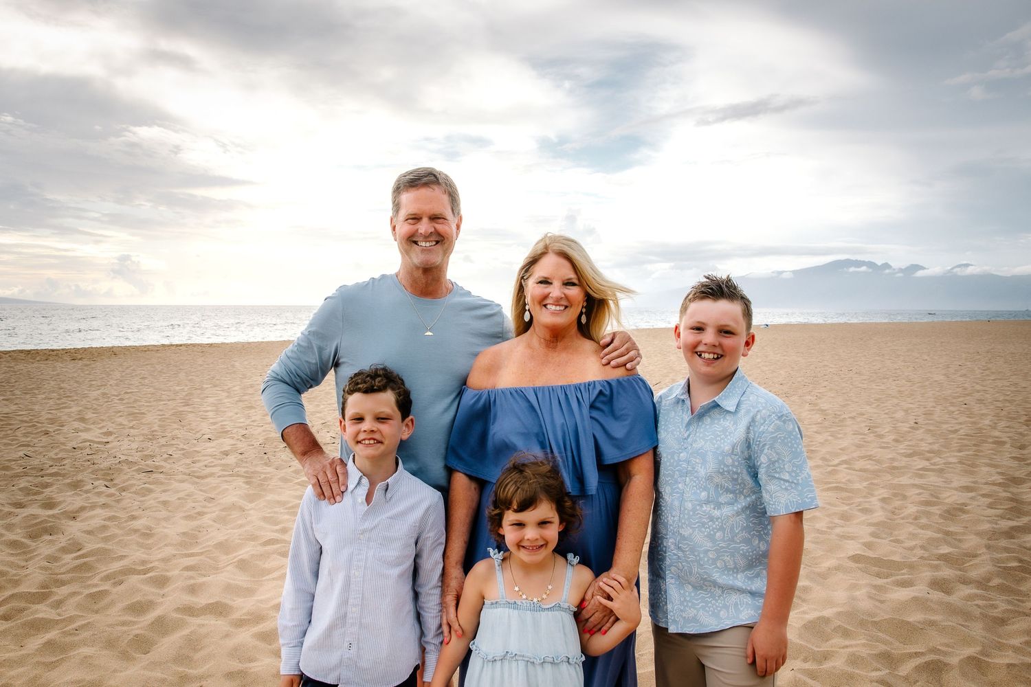 Family portrait session on a sandy beach with everyone wearing coordinated blue and white outfits.