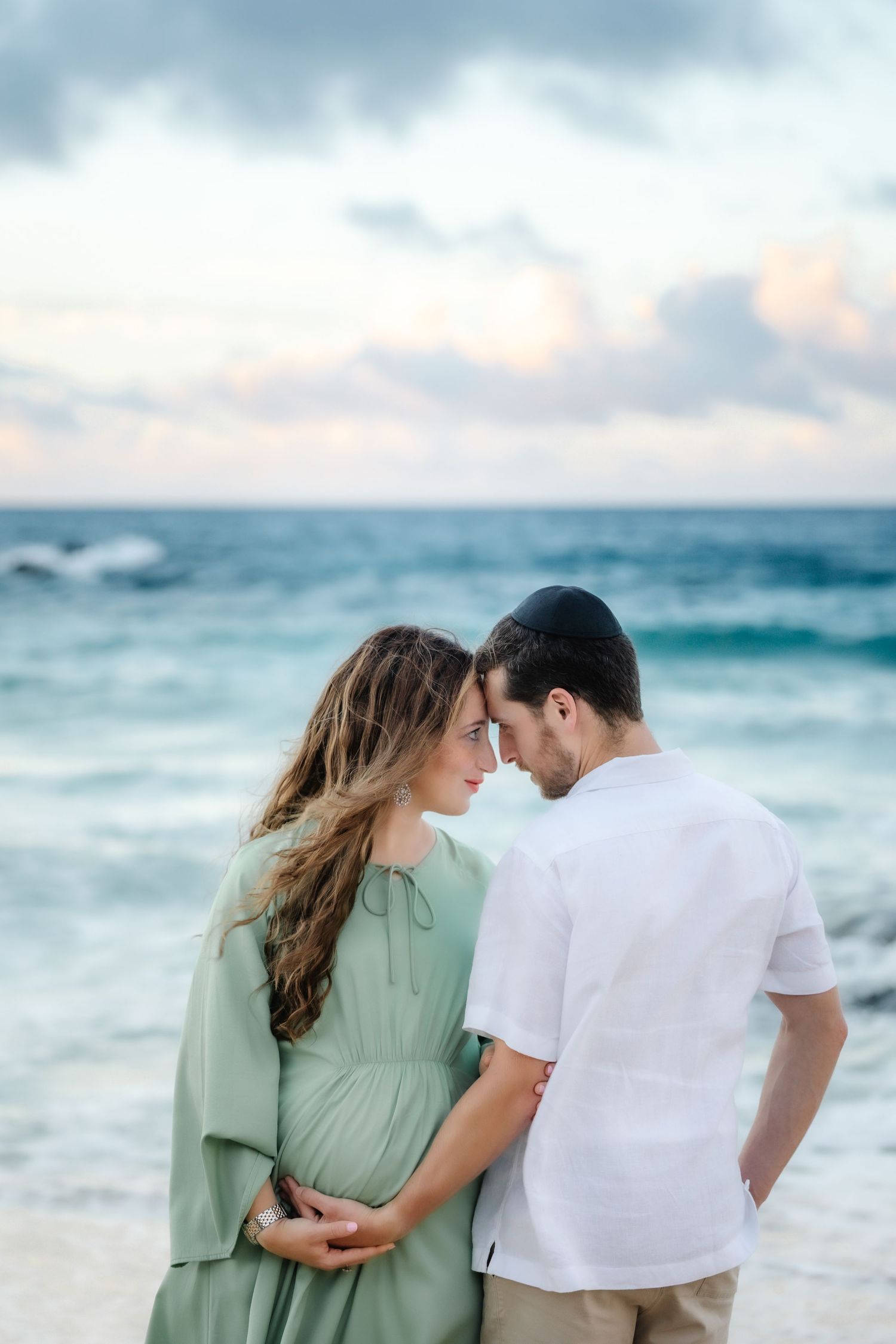 Pregnant couple sharing a tender moment on a beach at sunset, woman in sage green dress, man in white shirt.