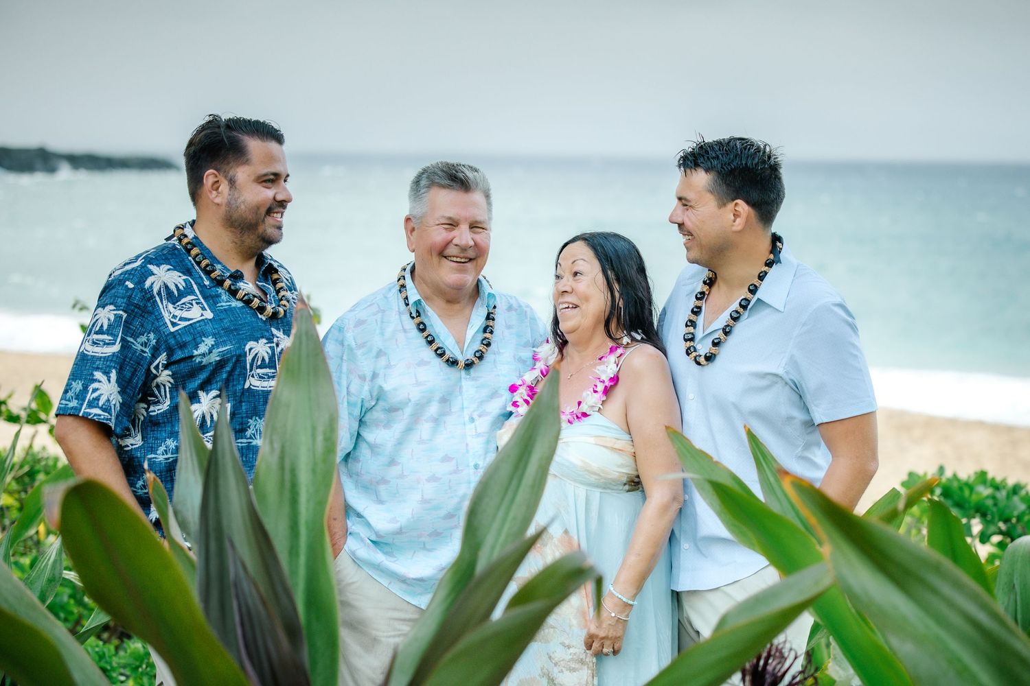 Four people laughing together on a tropical beach, wearing leis and Hawaiian attire, with ocean waves in the background.