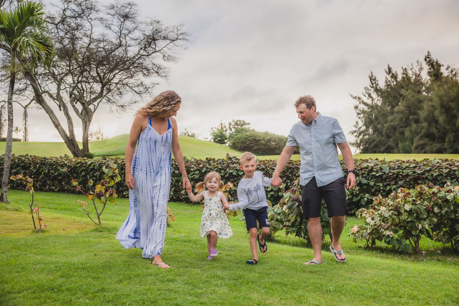 Family of four walking on green lawn, parents holding hands with two young children outdoors.