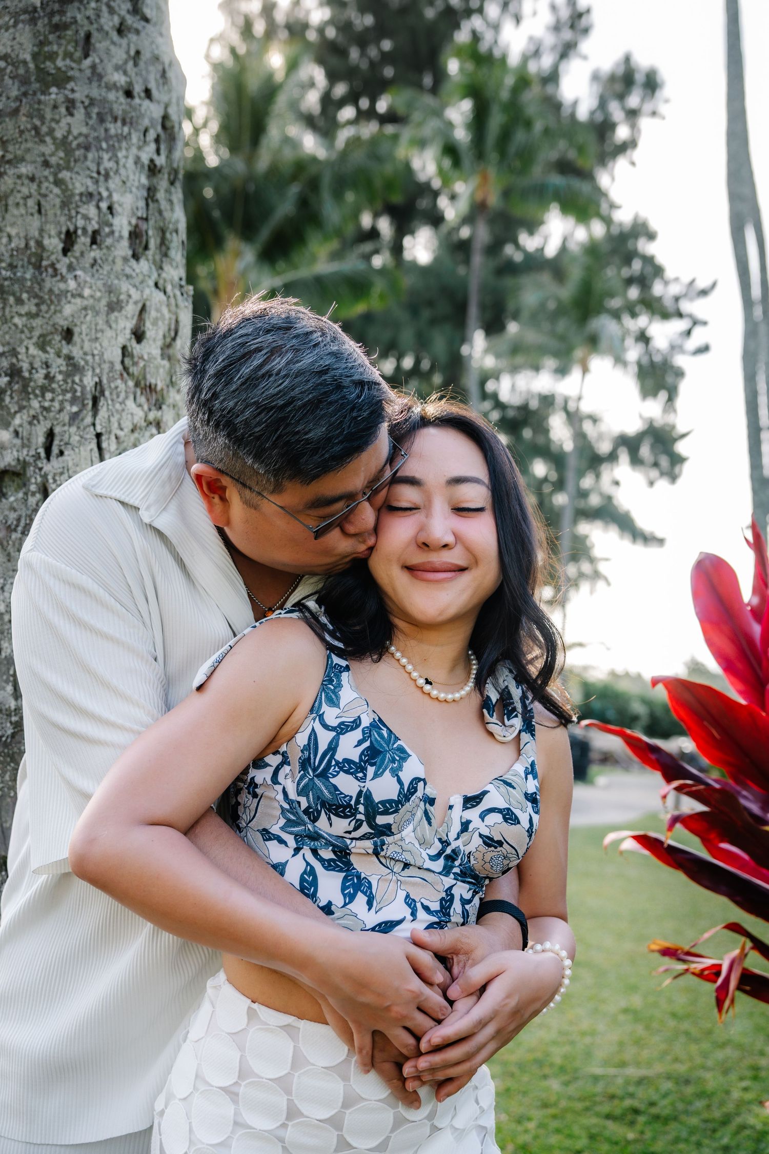Couple embracing near a palm tree in a tropical garden, man kissing woman's cheek as she smiles.