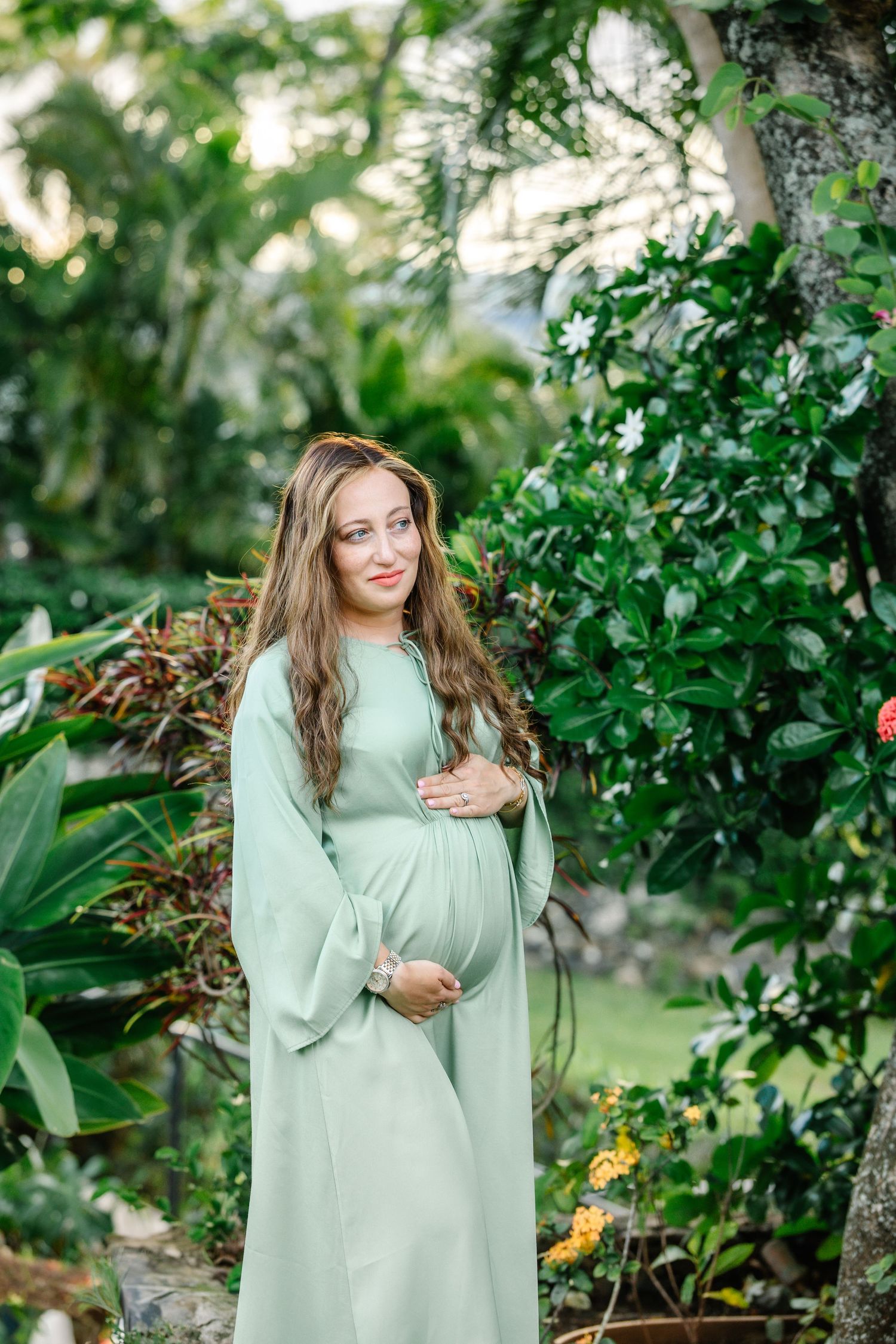 Pregnant woman in mint green dress cradling baby bump in lush tropical garden setting.