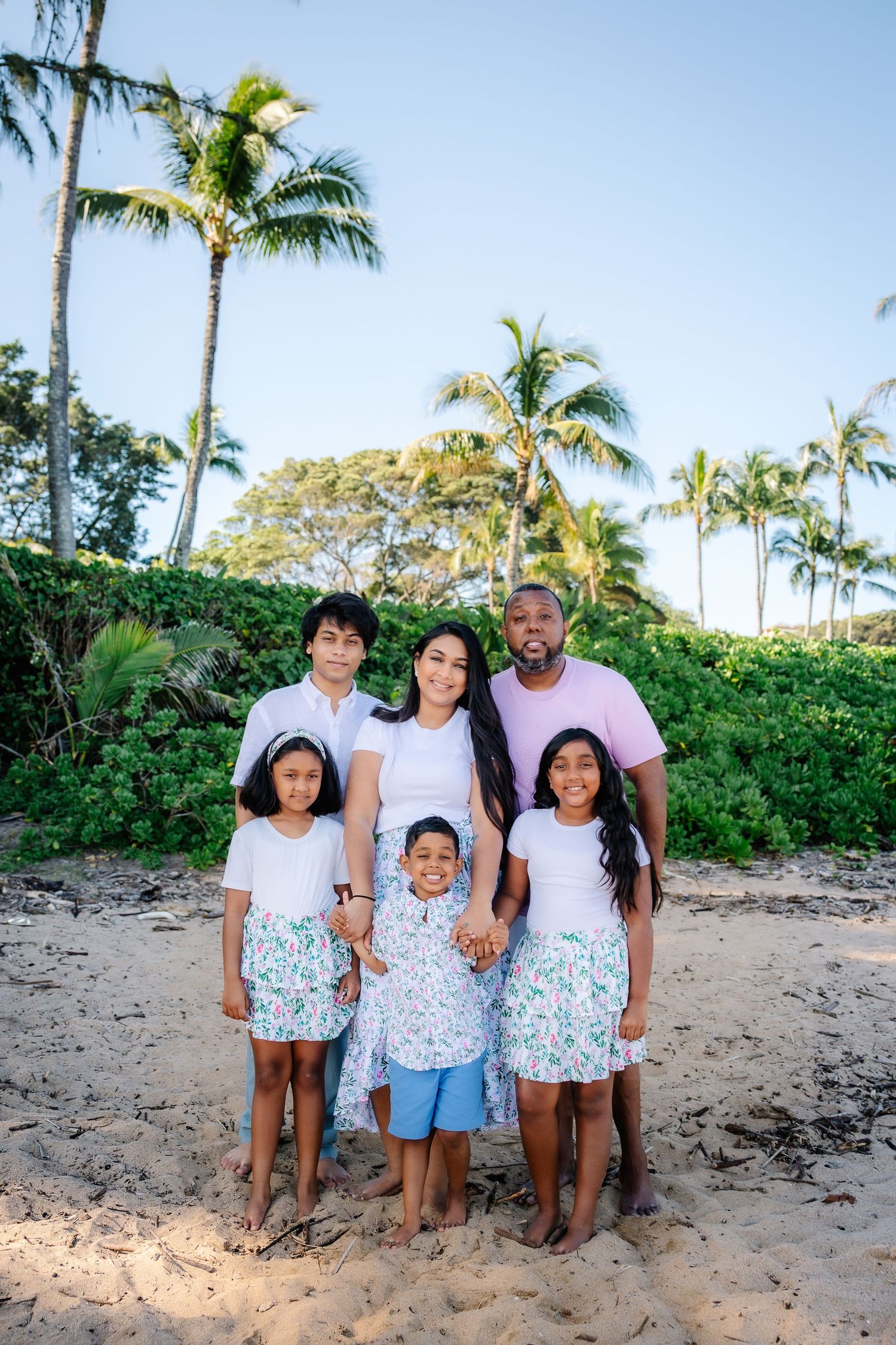 Family of six posing on sandy beach with palm trees, wearing coordinated white and floral outfits in a tropical setting.
