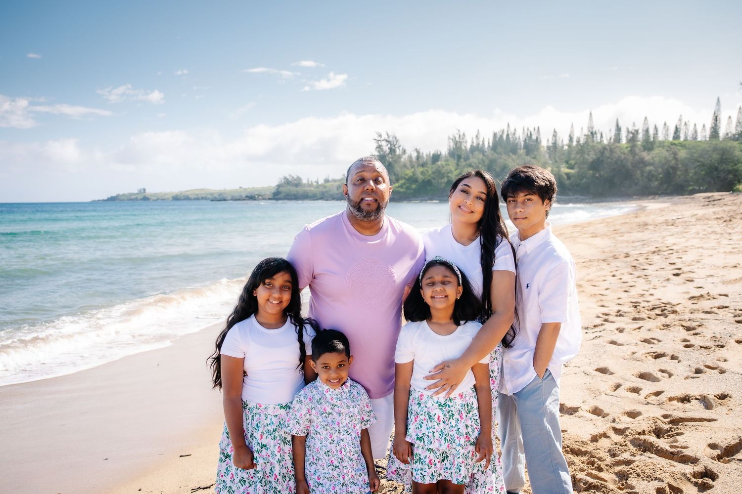 Family of six posing together on a sunny beach with turquoise ocean waves and lush green trees in the background.