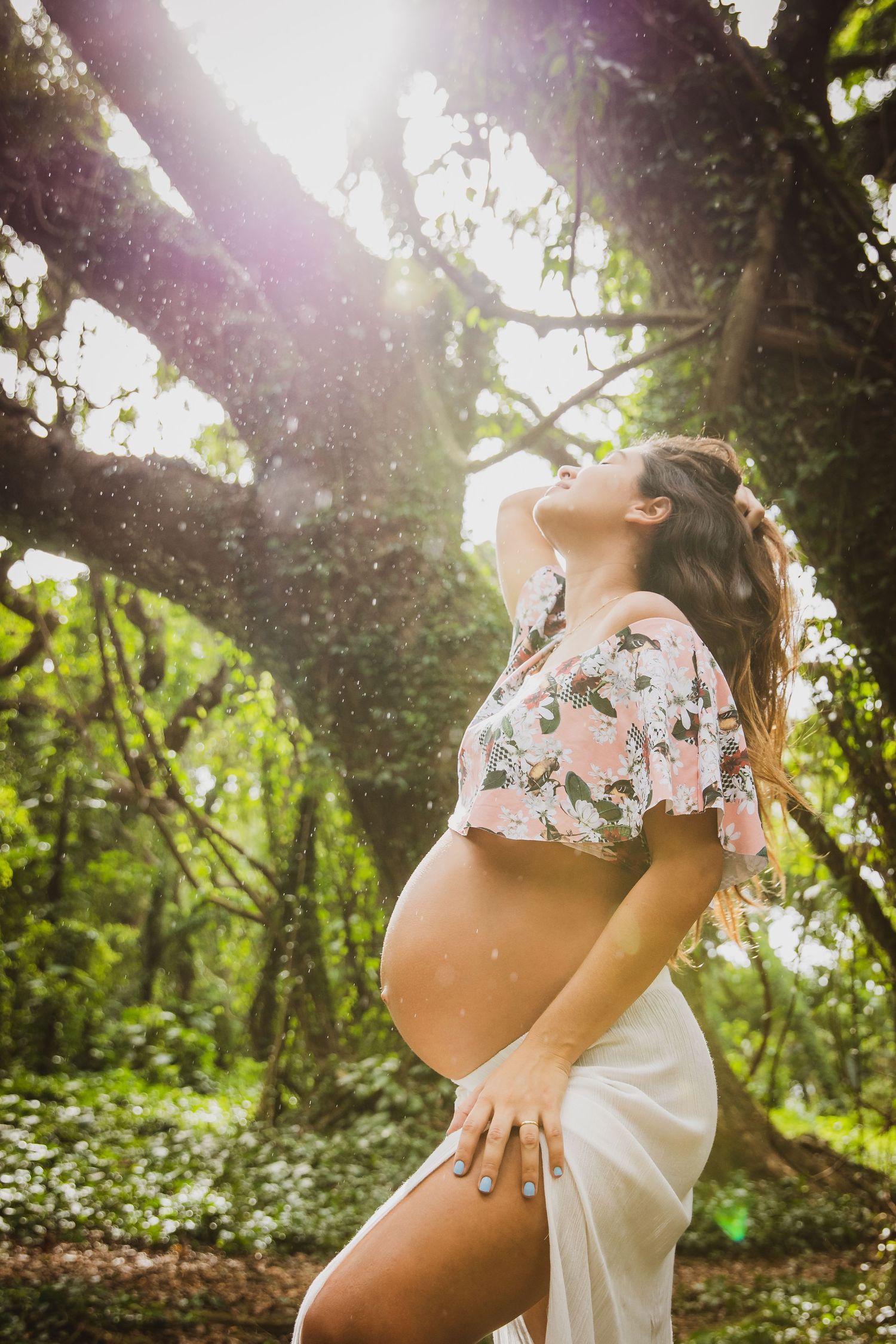 Pregnant woman in floral crop top poses outdoors under large tree with sunlight streaming through lush green foliage.