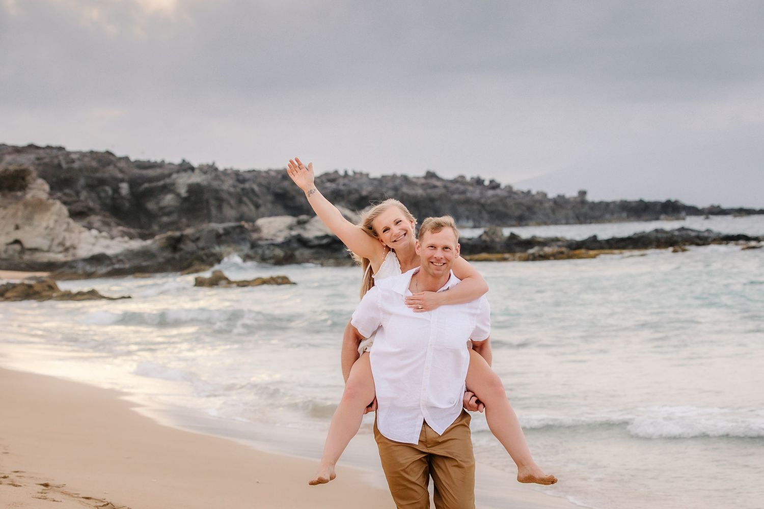 Couple enjoying a piggyback ride on a sandy beach, woman raising her arm joyfully with ocean waves and rocks behind.