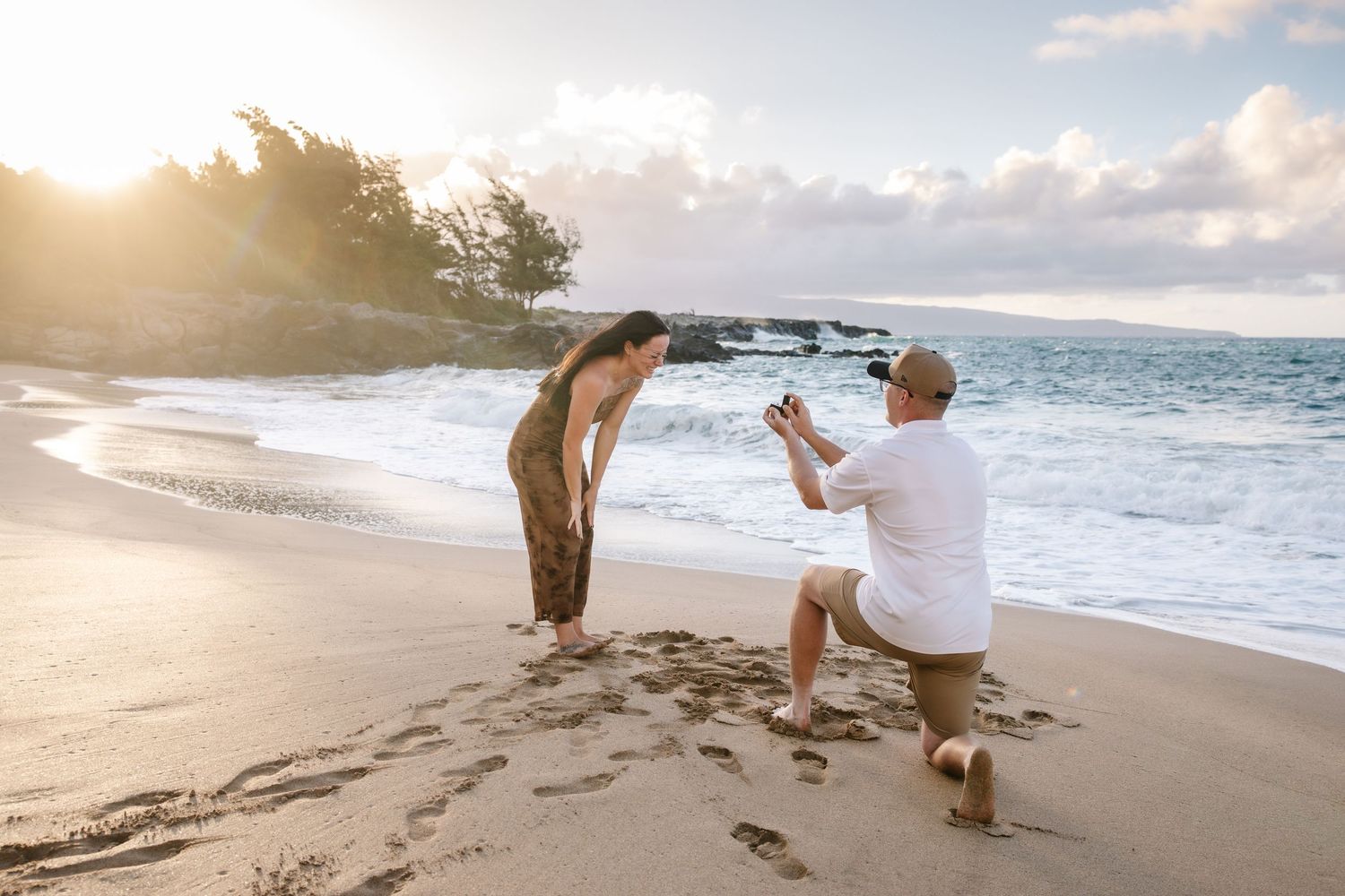 Man proposes on a sunny beach at sunset while woman leans in to see, waves and golden light in background.