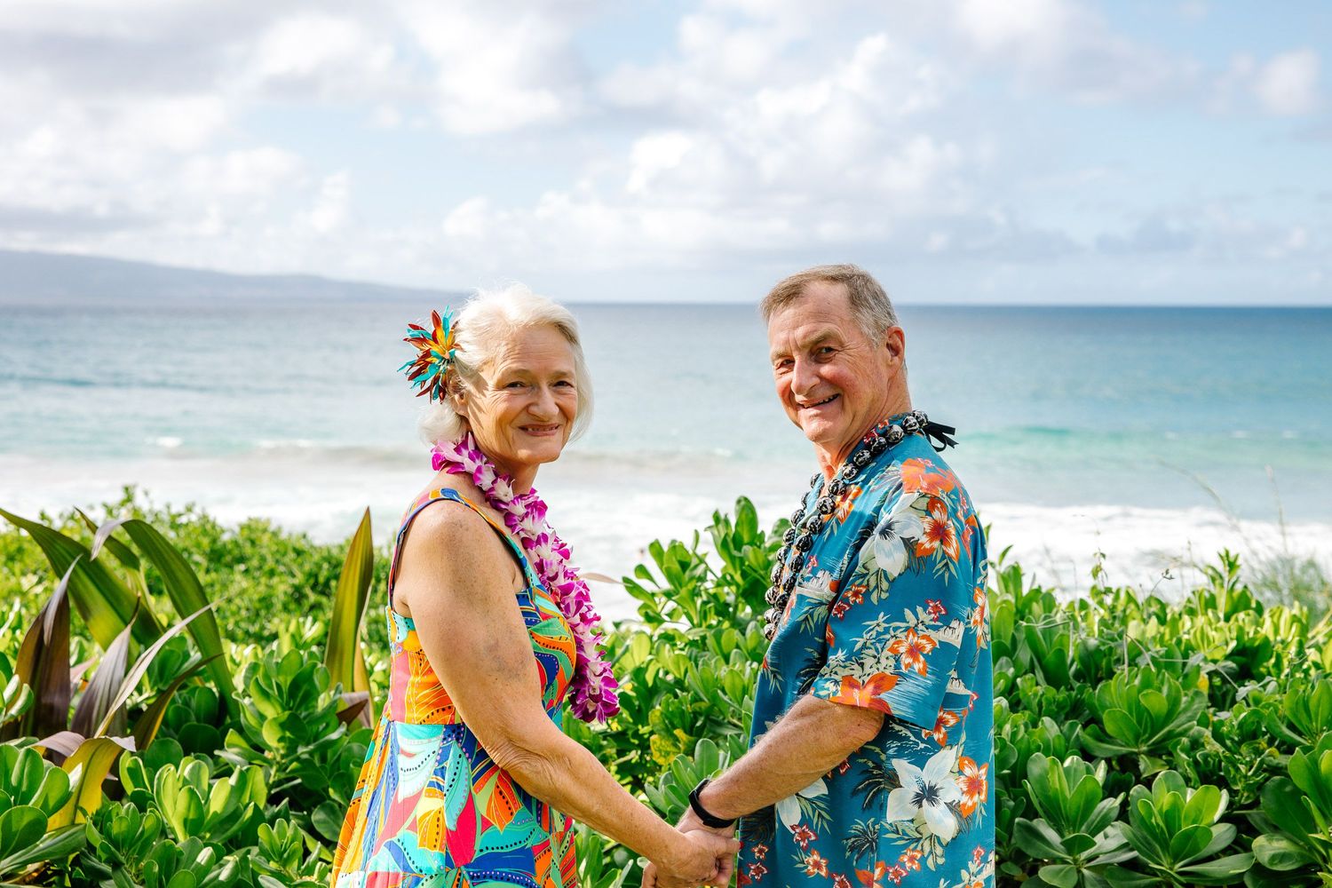 Couple in matching Hawaiian print outfits posing together on a tropical beach with lush greenery.