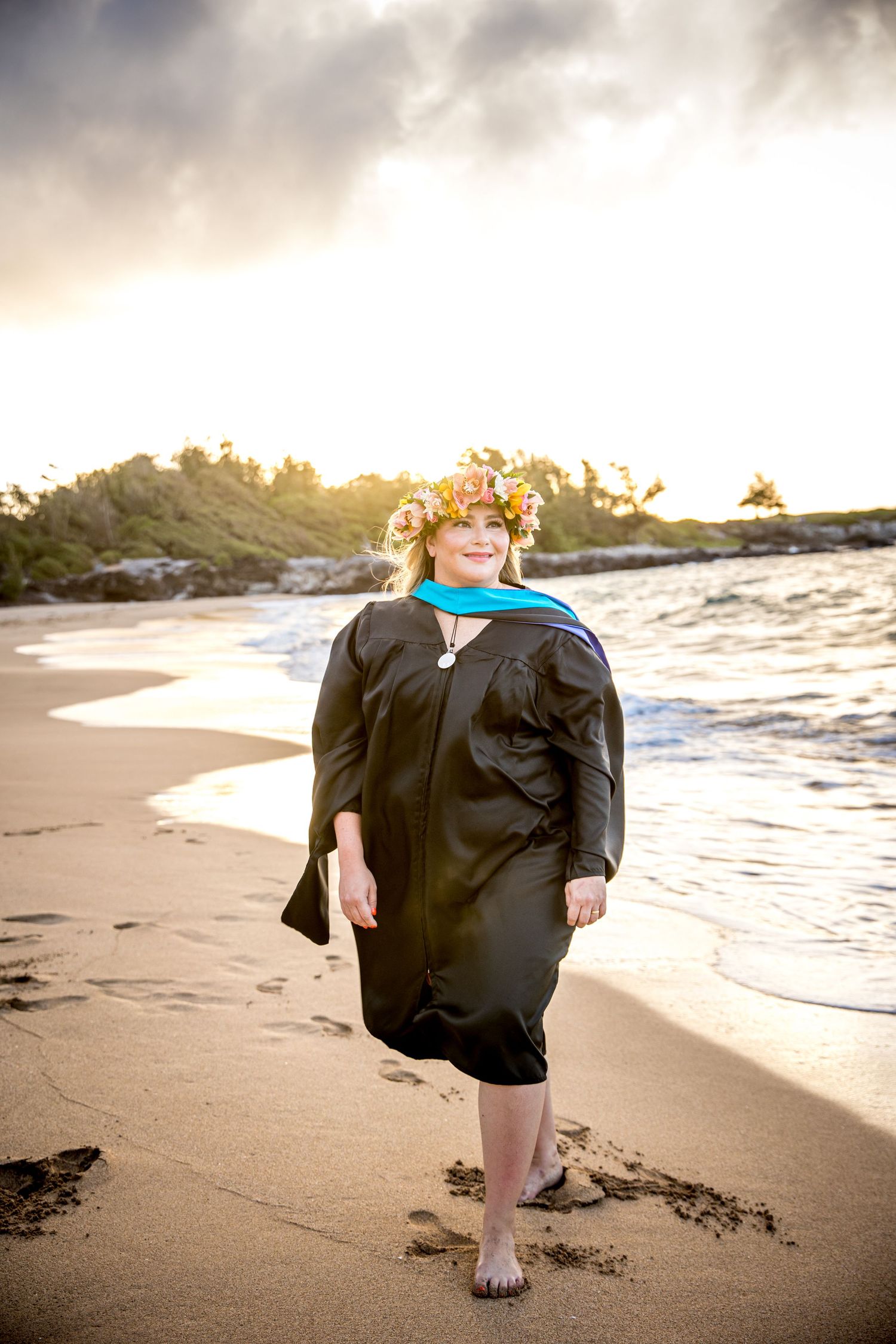 Graduation photoshoot  woman walks along a sandy beach at sunset with dramatic cloudy skies overhead.
