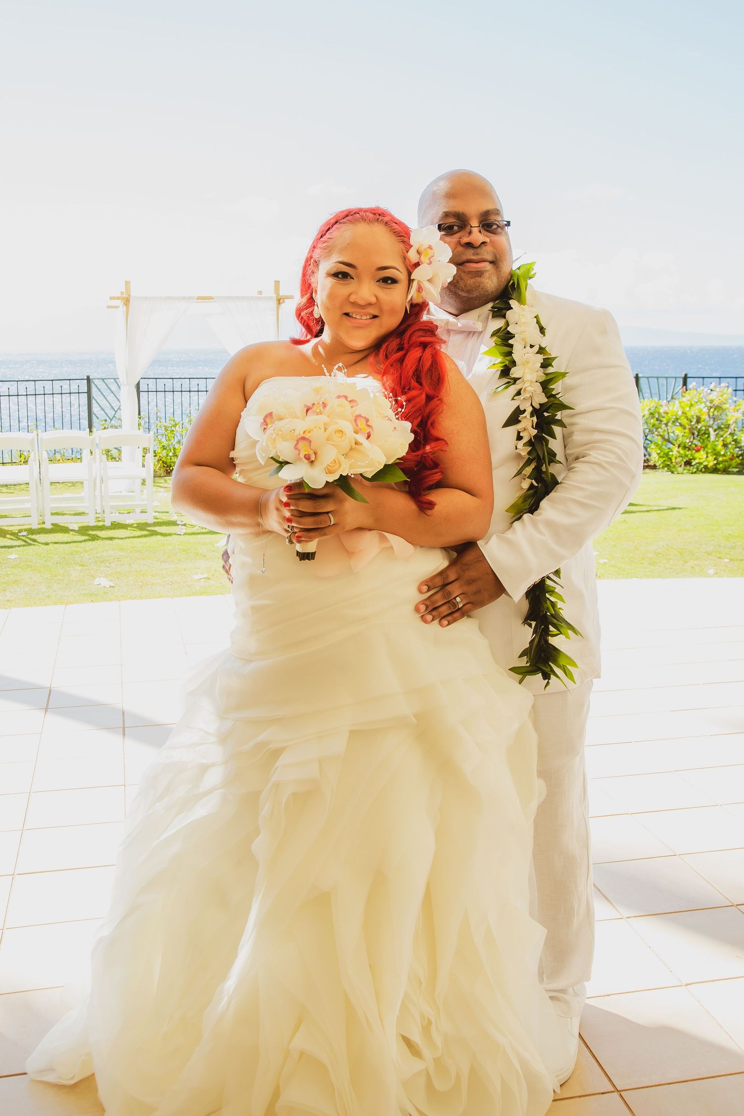 Bride with red hair holding white bouquet poses with groom wearing lei in white suit at outdoor ocean wedding.