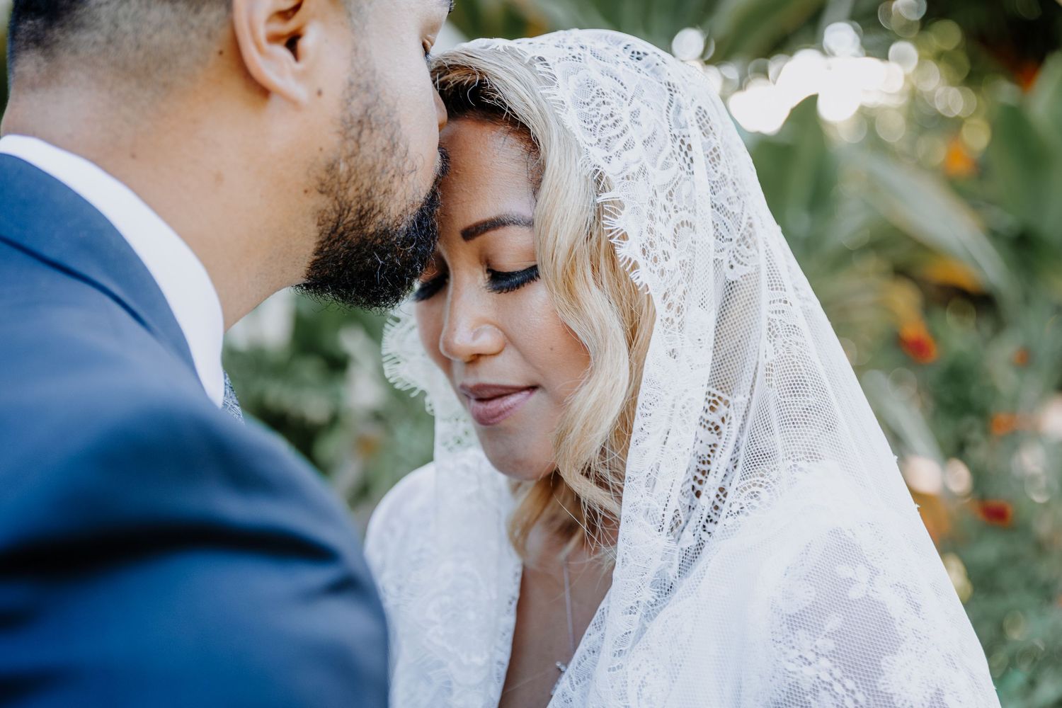 A bride in a lace veil and groom in a blue suit share an intimate moment outdoors surrounded by greenery.